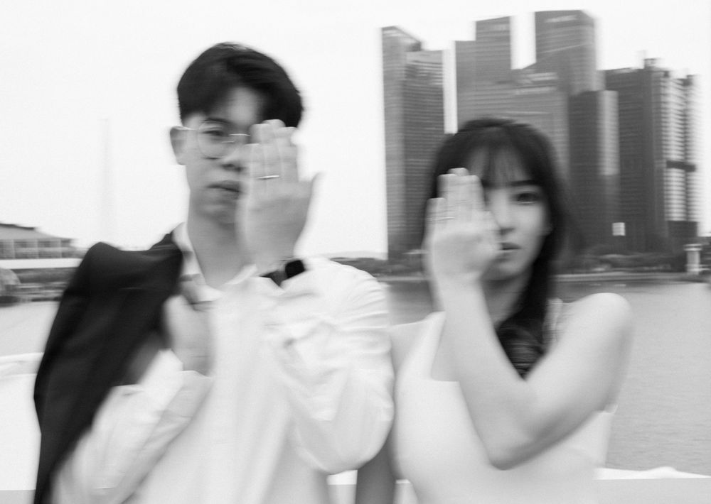 Black and white photo of a young Asian couple blocking camera with hands, city skyline in background.