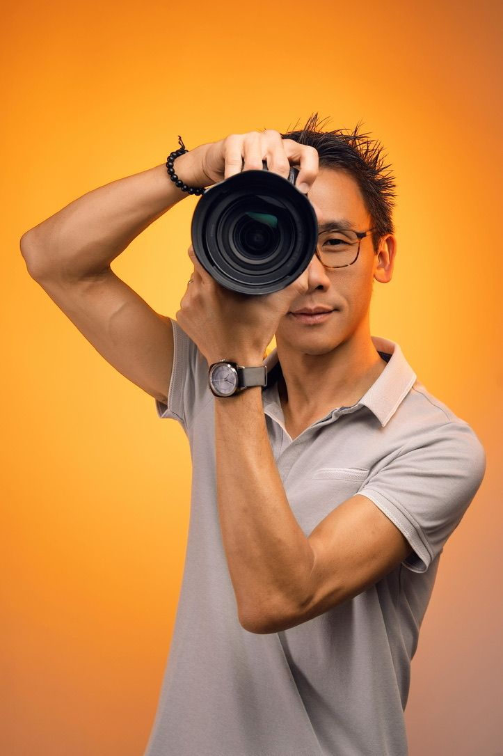 Smiling male photographer holding DSLR camera with telephoto lens overhead, wearing glasses and polo shirt on orange background.