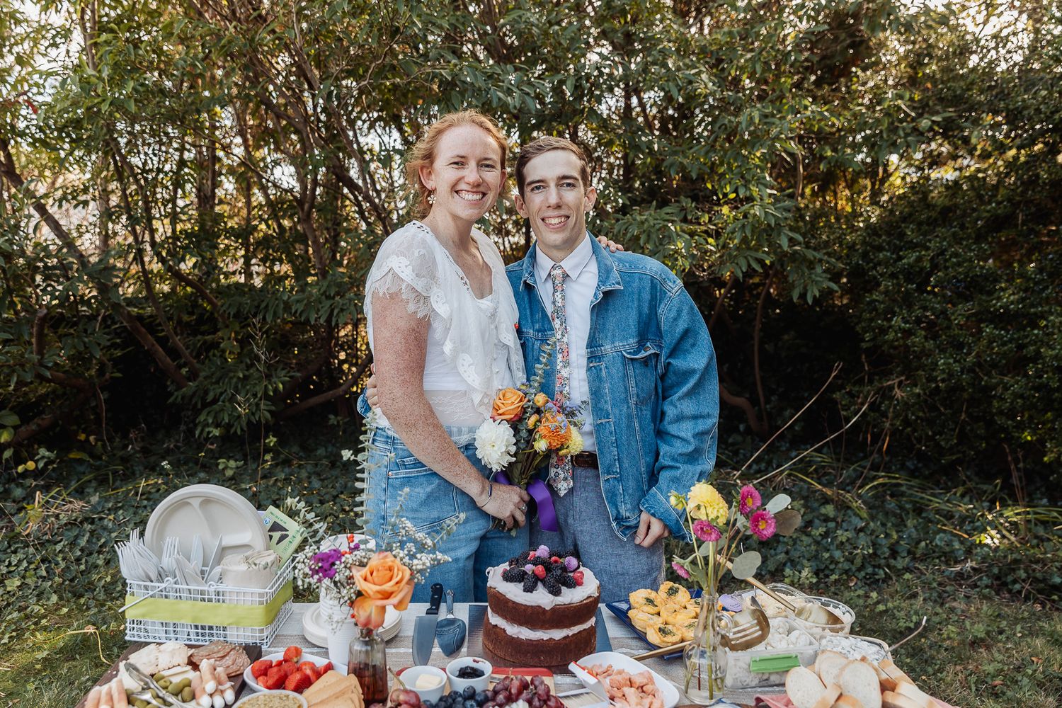 Smiling couple in casual wedding attire standing behind a colorful dessert table outdoors surrounded by lush greenery.