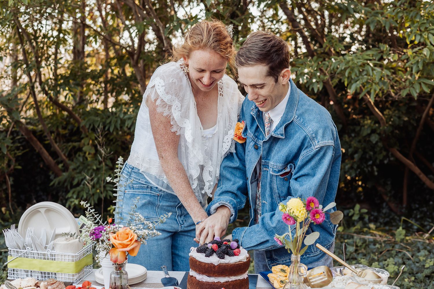 Couple cutting a naked wedding cake outdoors, surrounded by colorful flowers and greenery, wearing casual denim attire.