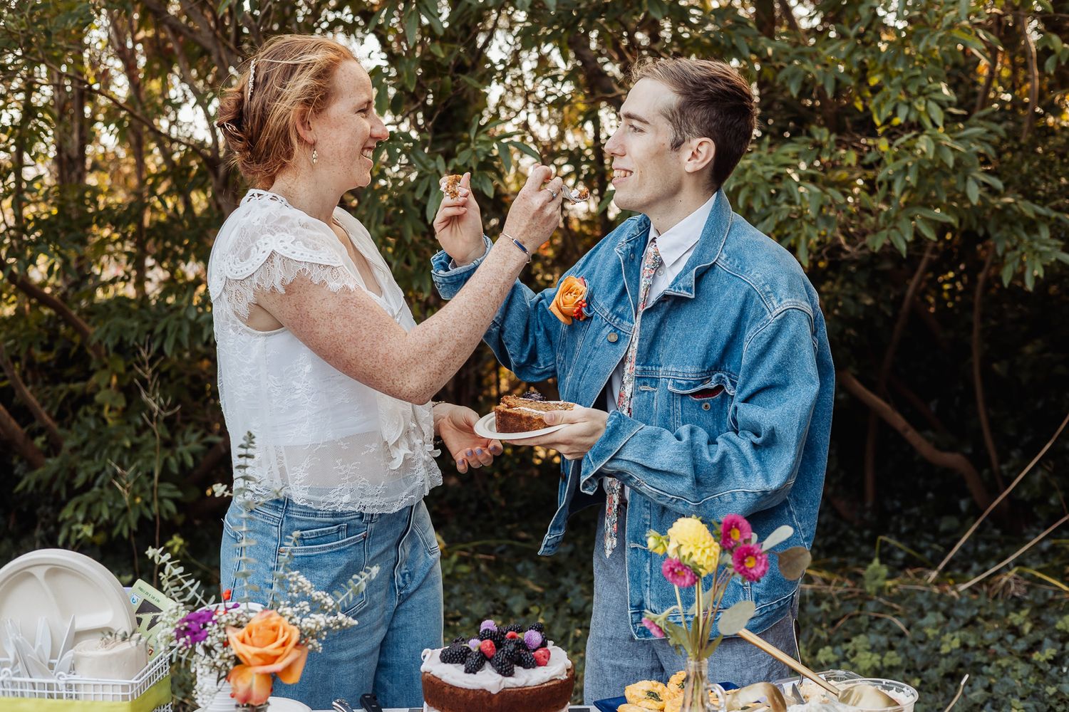 Two brides share cake outdoors at a colorful garden wedding celebration, dressed in denim and white lace.