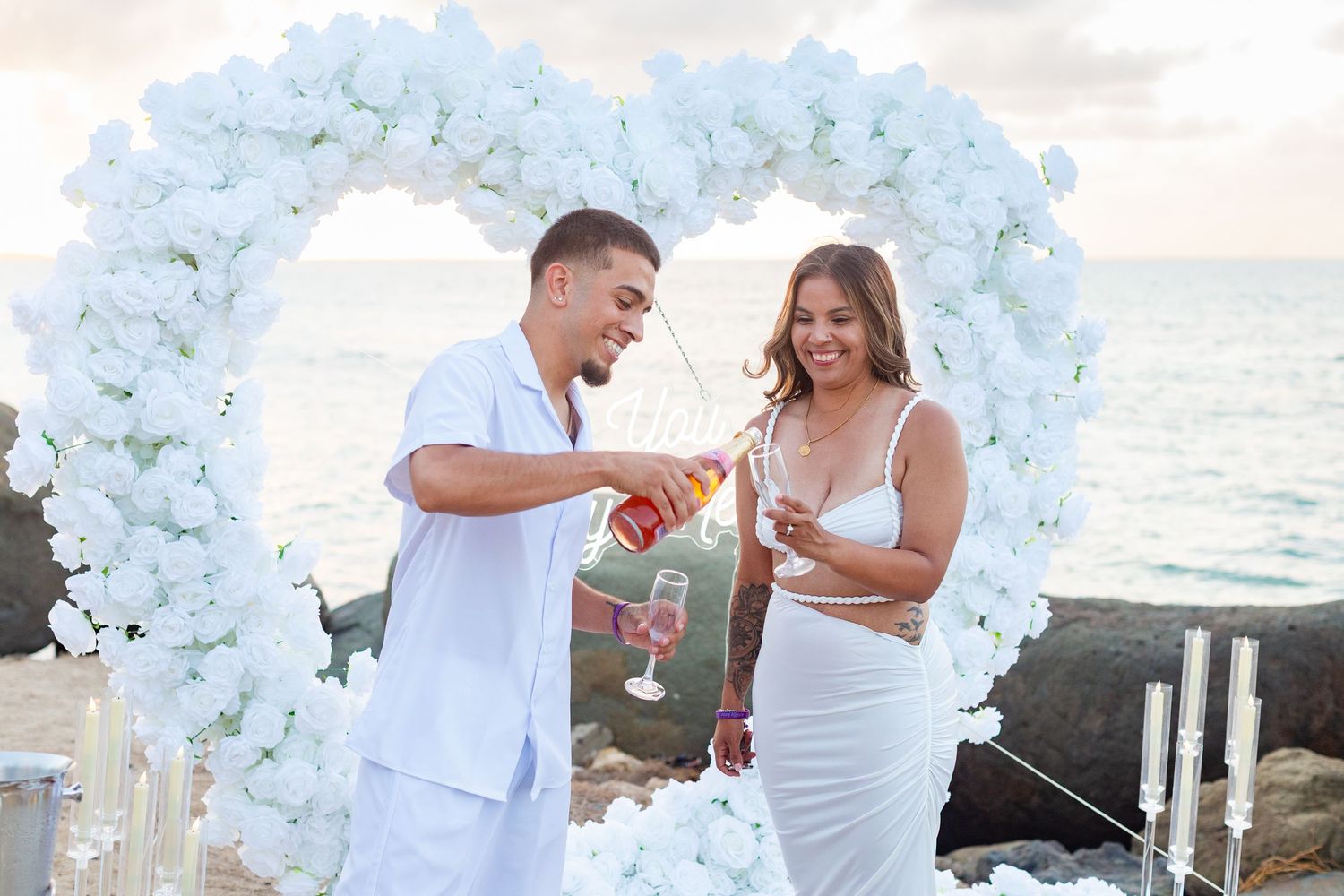 Marriage proposal. Toast. Man, woman, and decorative setup with white flower heart overlooking the ocean in Río Grande, Puerto Rico.
