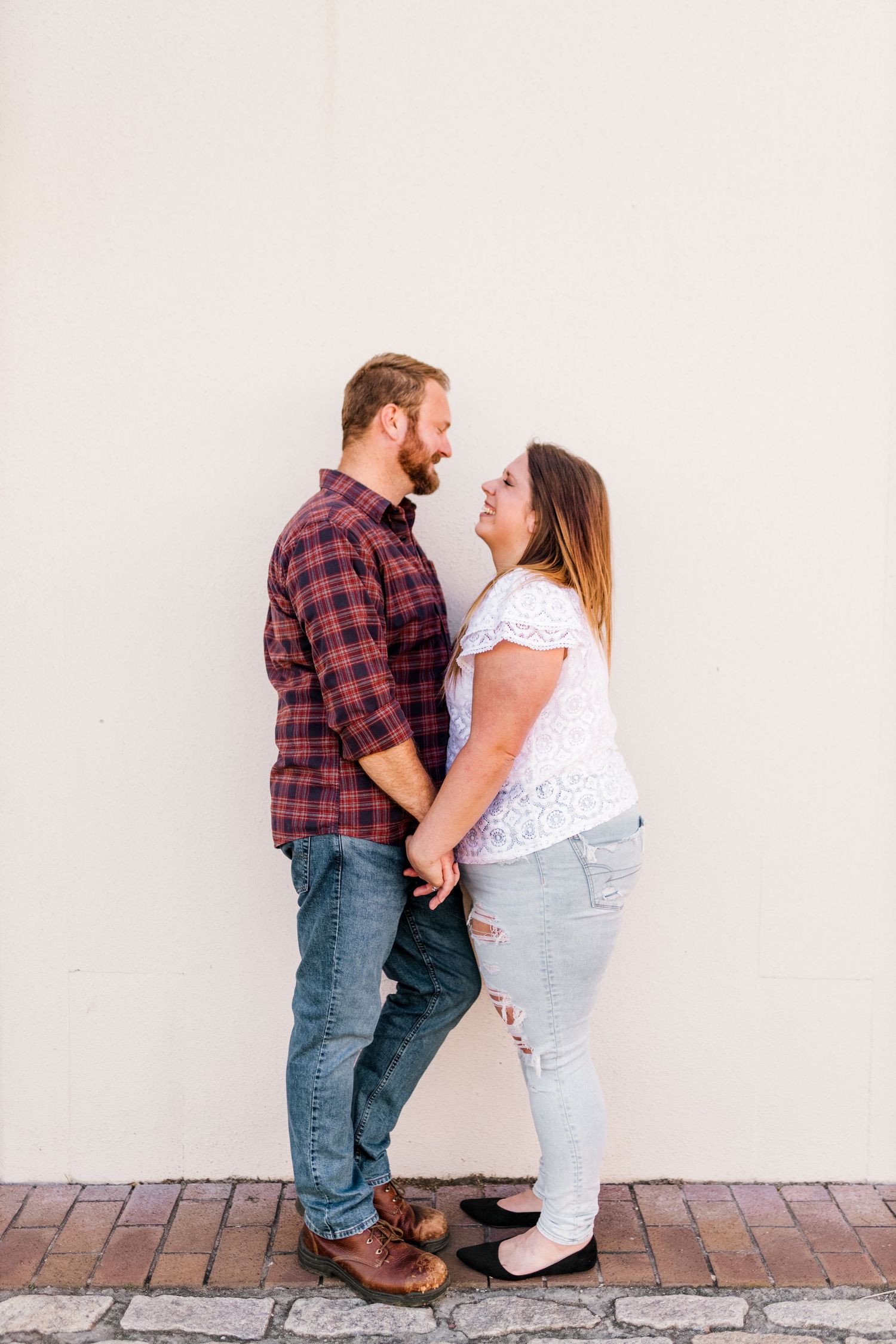 A couple holds hands and gazes at each other against a white wall, wearing casual jeans and plaid shirt.