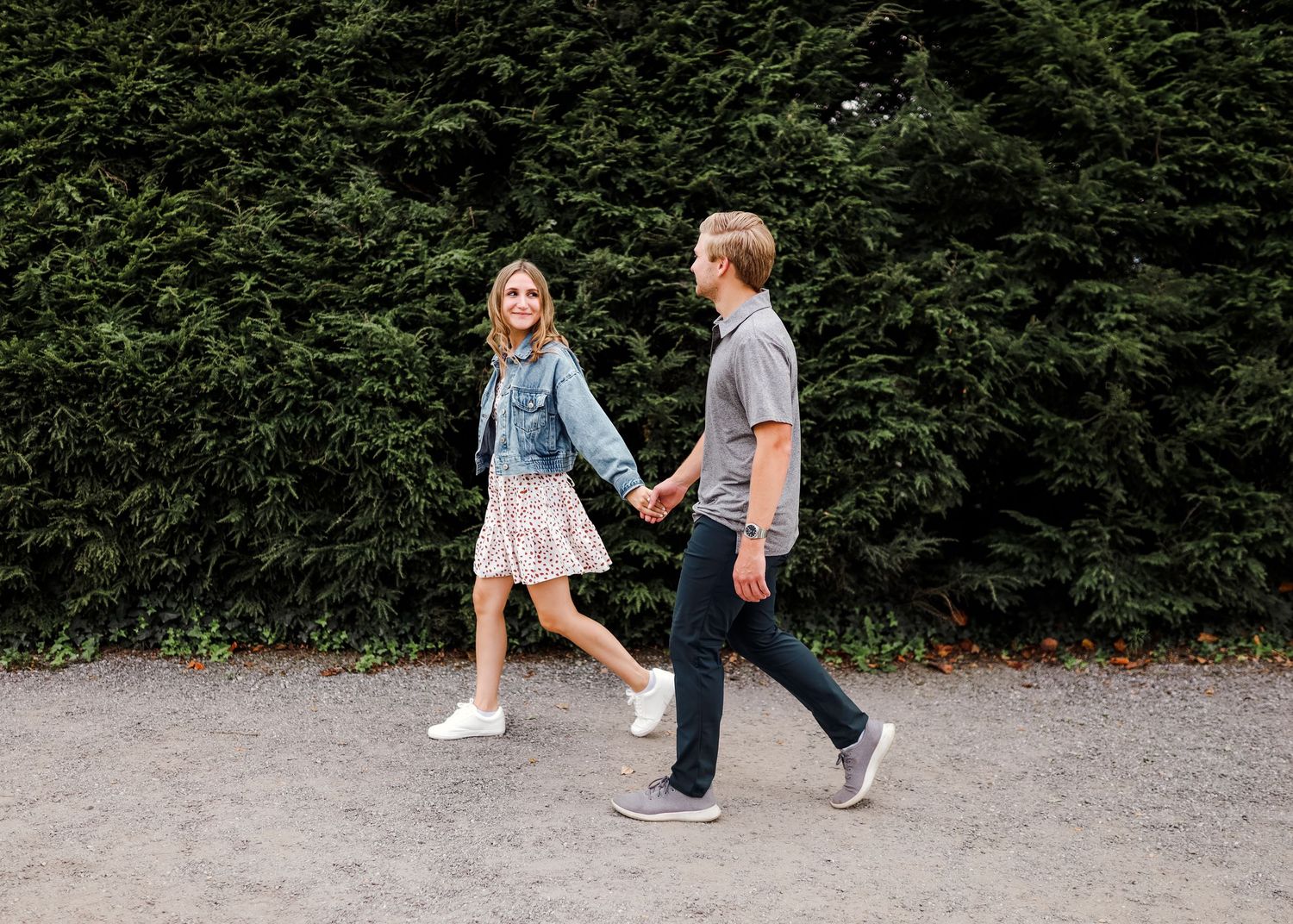 A young couple walks hand-in-hand on a gravel path in front of a lush green hedge wall.