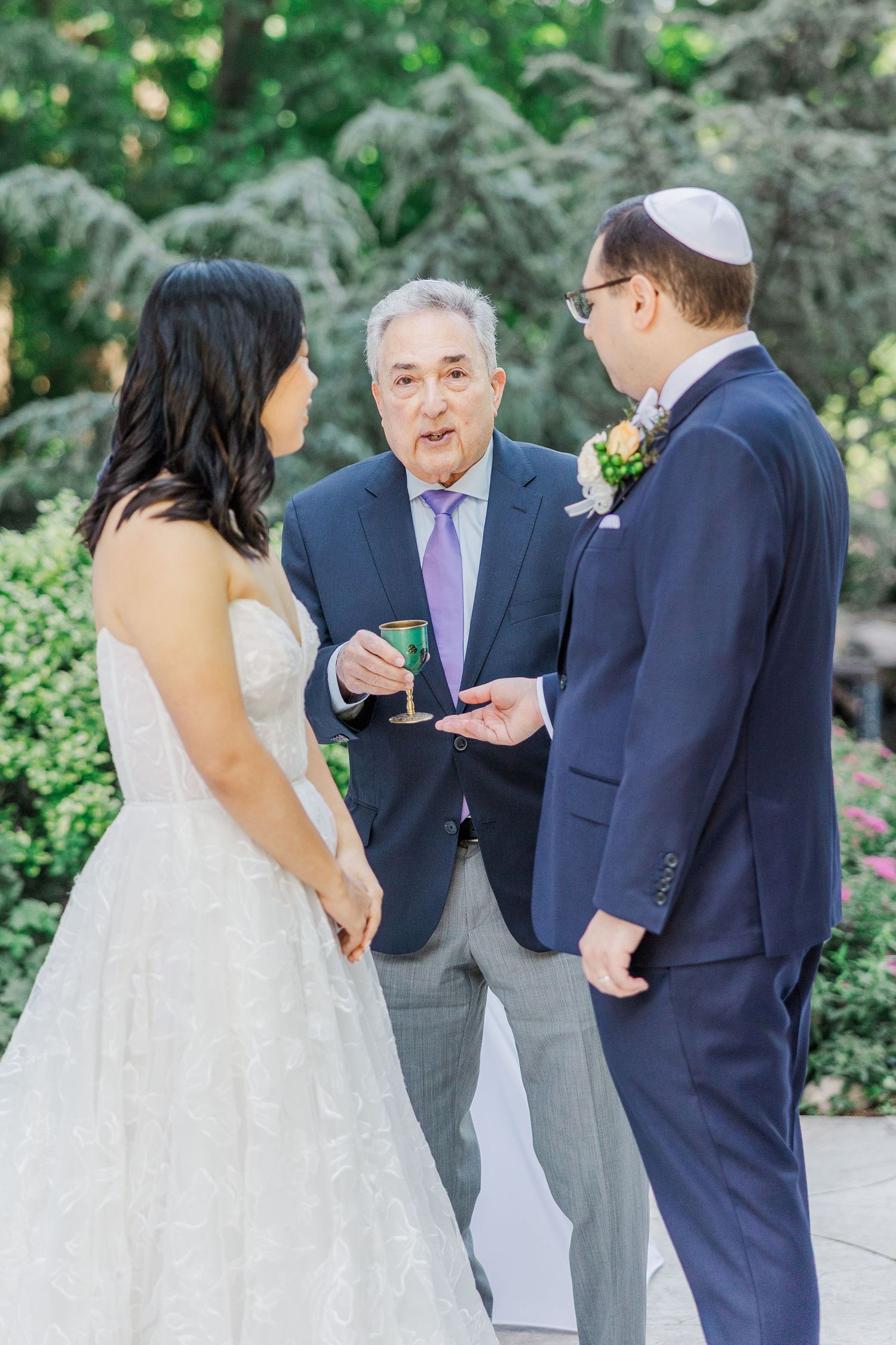 Rabbi officiating outdoor Jewish wedding ceremony with bride and groom, holding ceremonial cup amid lush garden backdrop.
