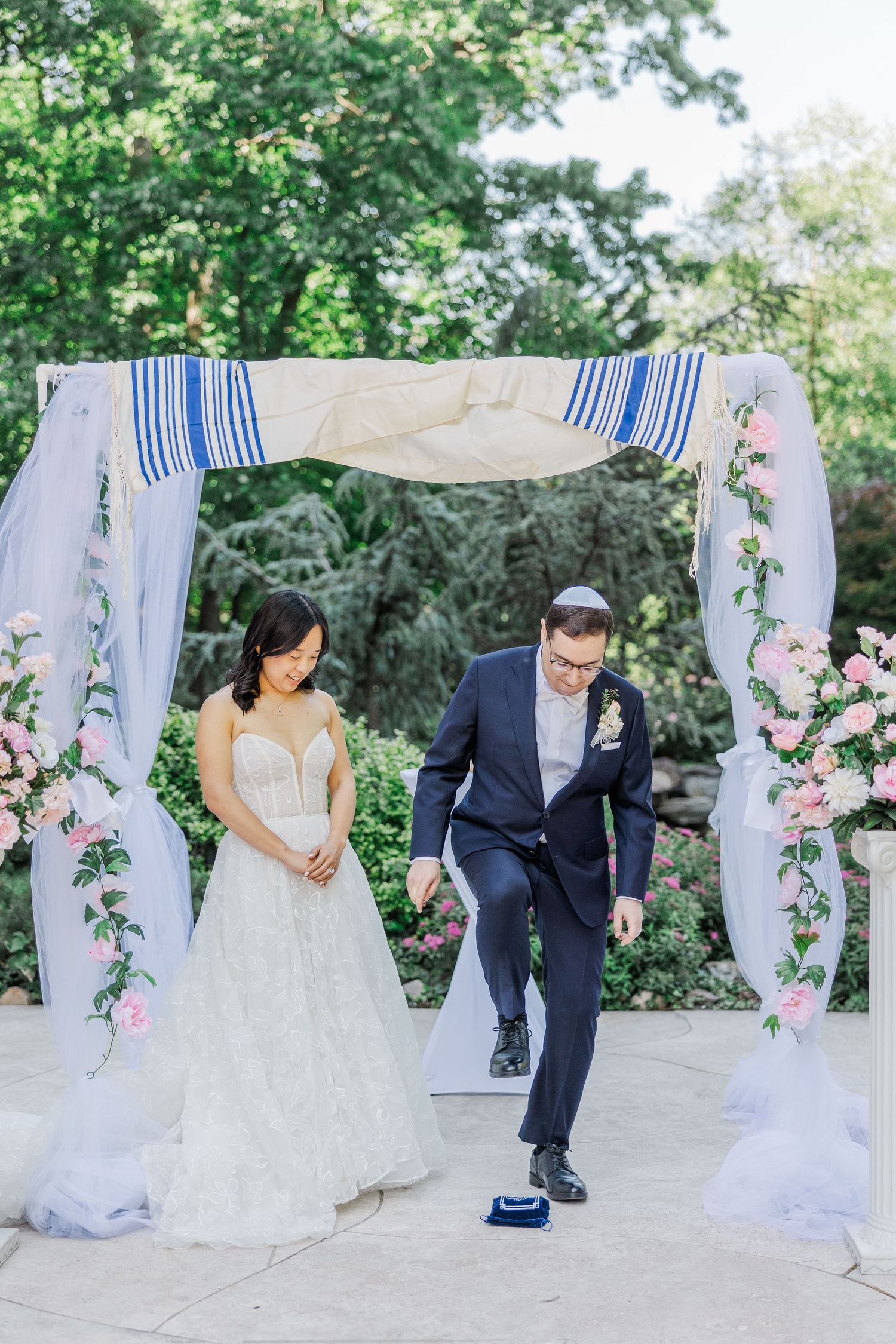 Groom stomping glass under floral chuppah at outdoor Jewish wedding ceremony with bride in strapless gown.