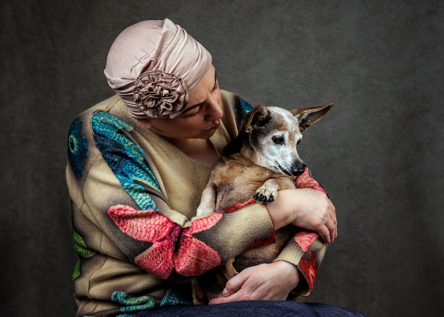 Elderly woman in pink turban lovingly embracing a small brown dog against a dark studio background.