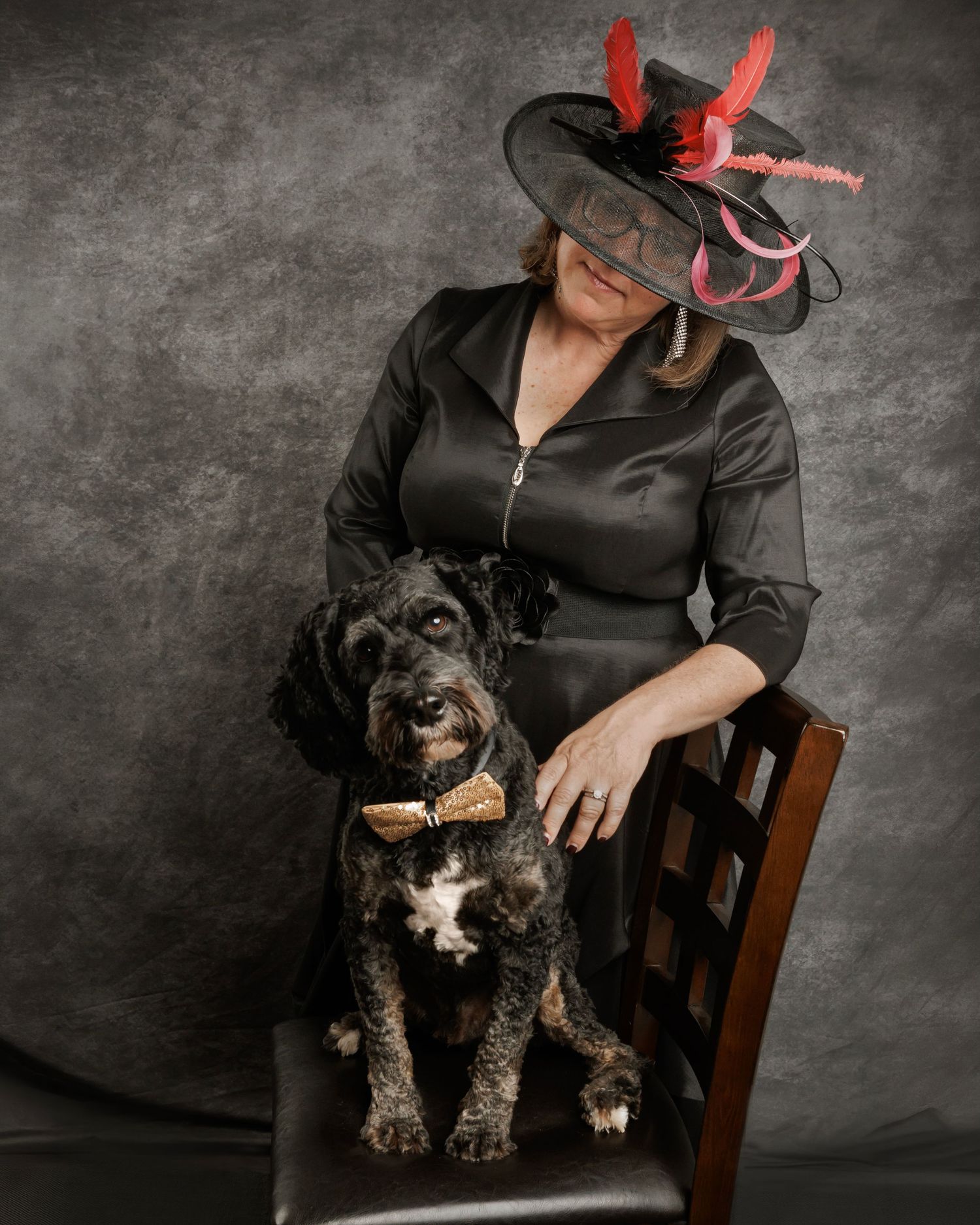Woman in black dress and red-feathered hat poses with a black curly dog wearing a gold bow tie on a chair.