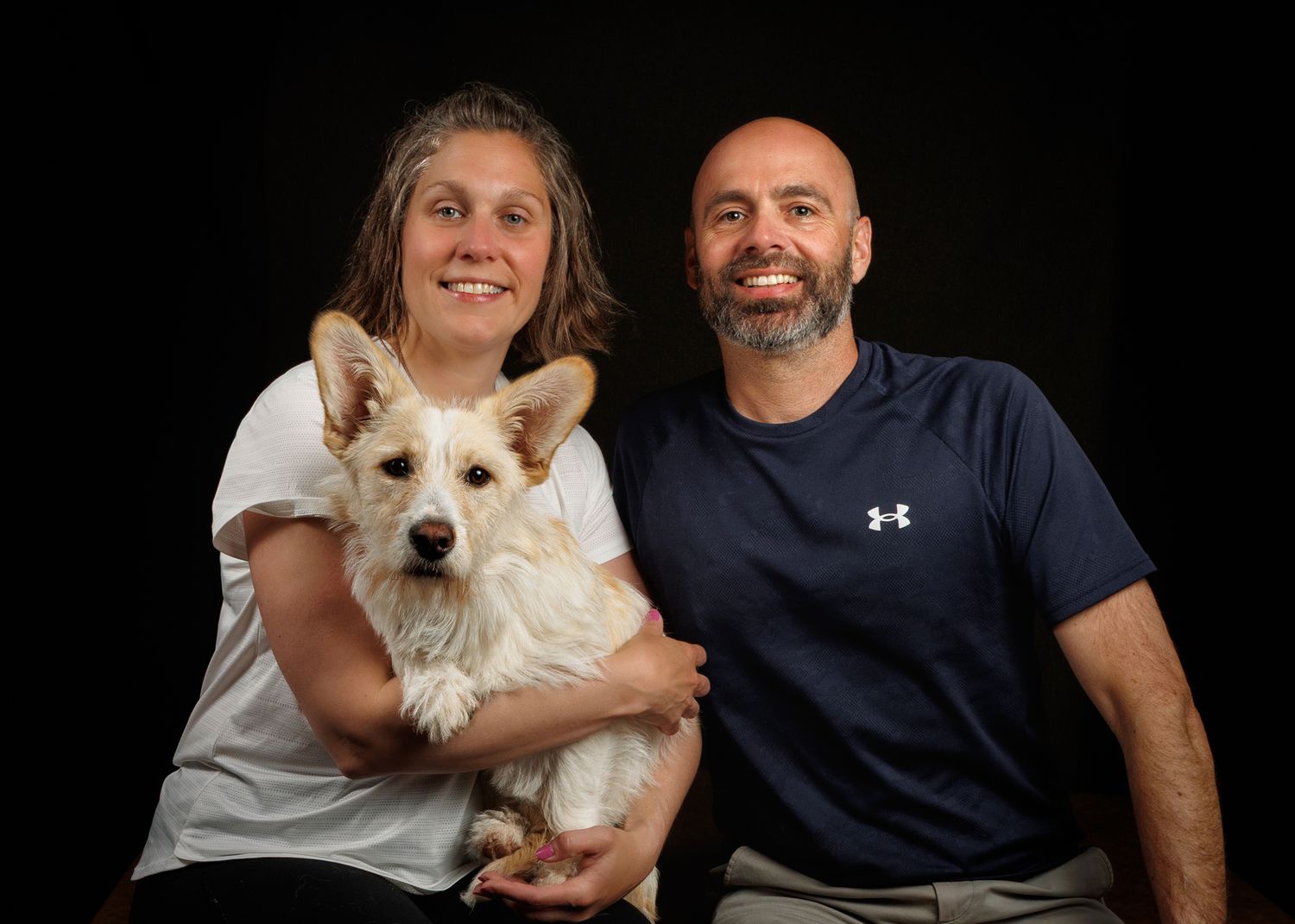 A smiling couple poses with a fluffy white dog against a dark background in a professional portrait photo.