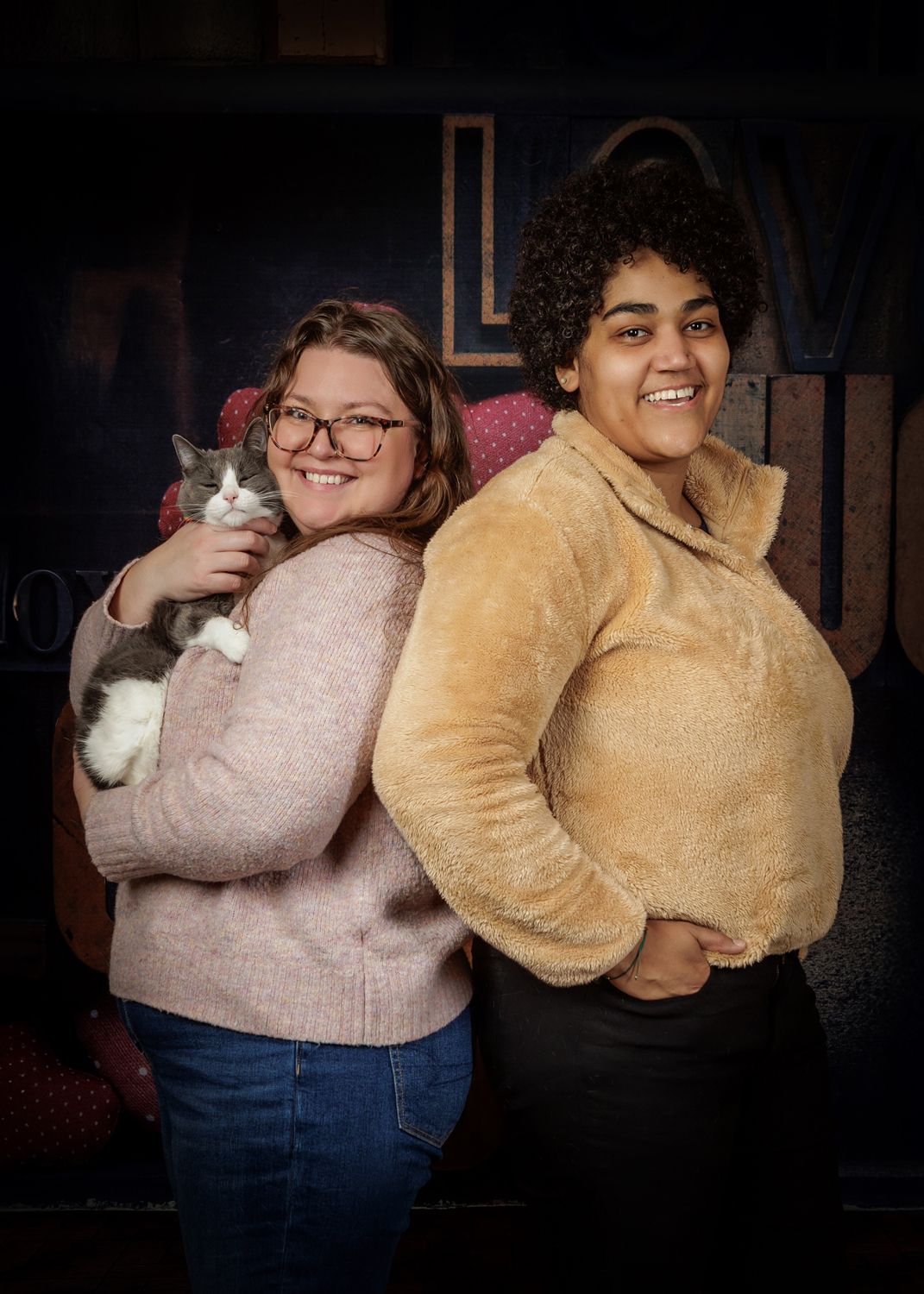 Two young women smiling and posing back-to-back, one holding a gray and white cat, in front of a decorative backdrop.