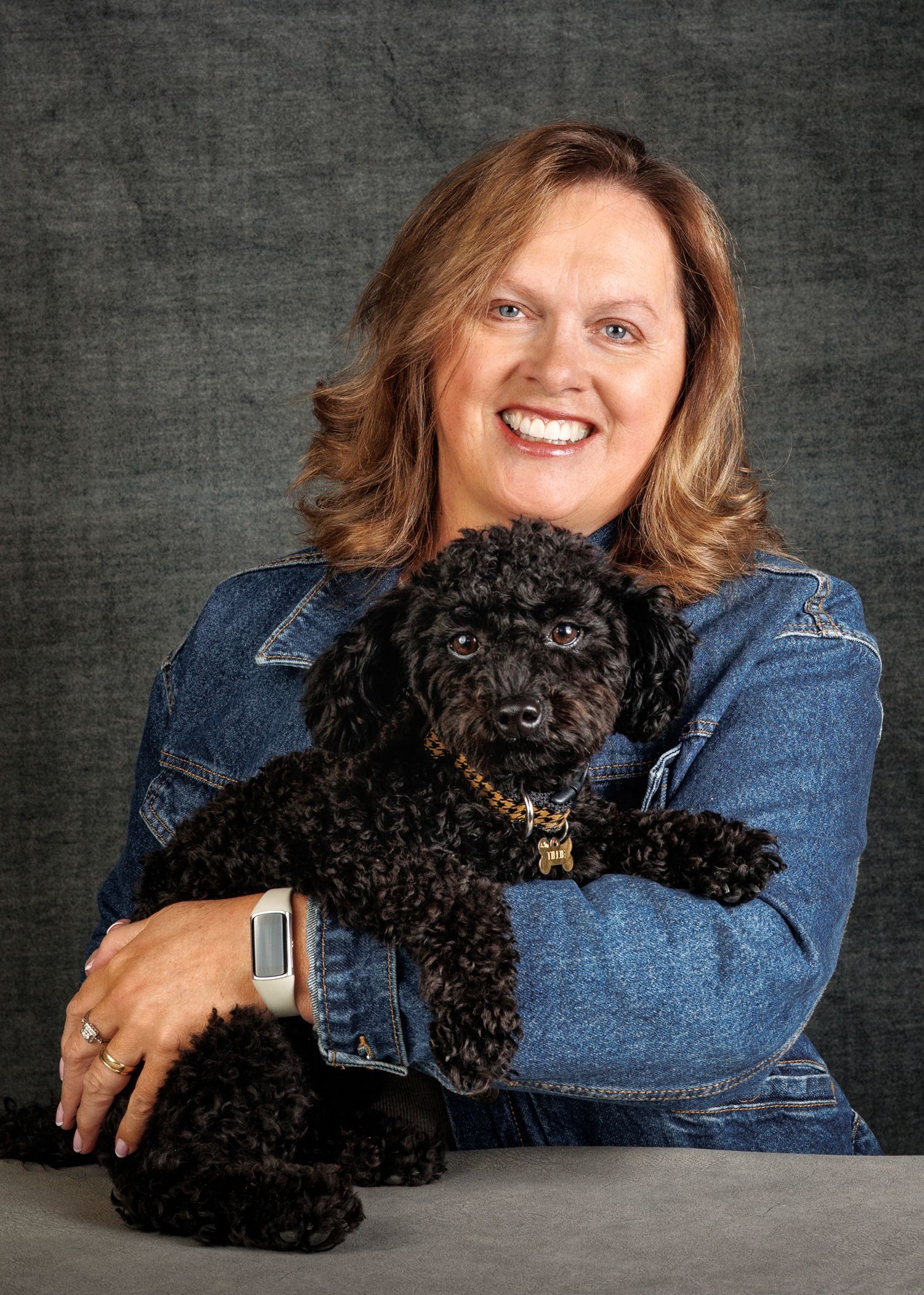 Smiling woman in denim jacket holding a small black curly-haired poodle puppy against a gray studio background.