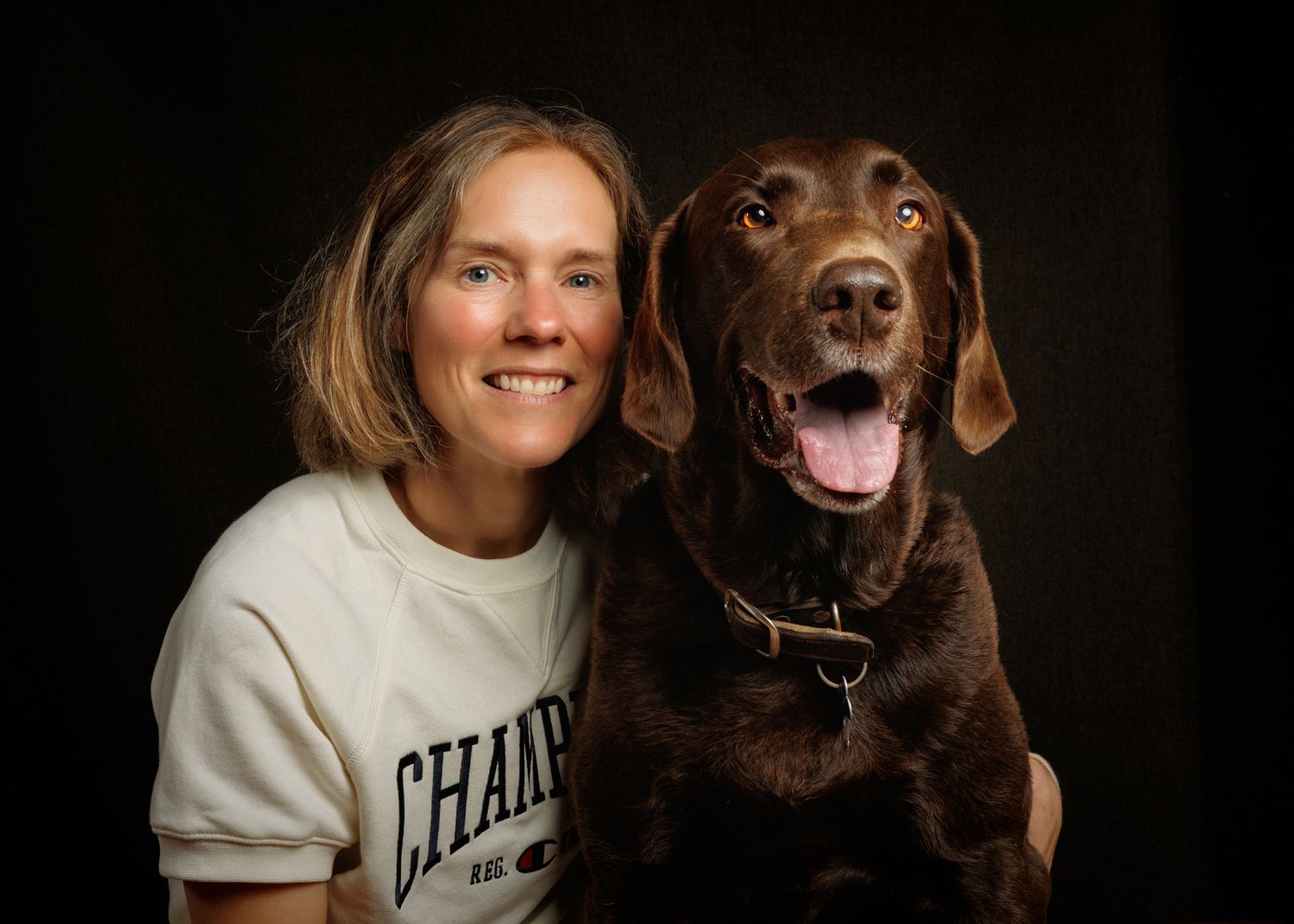 A smiling woman in a Champion sweatshirt poses with a happy chocolate Labrador Retriever against a dark background.