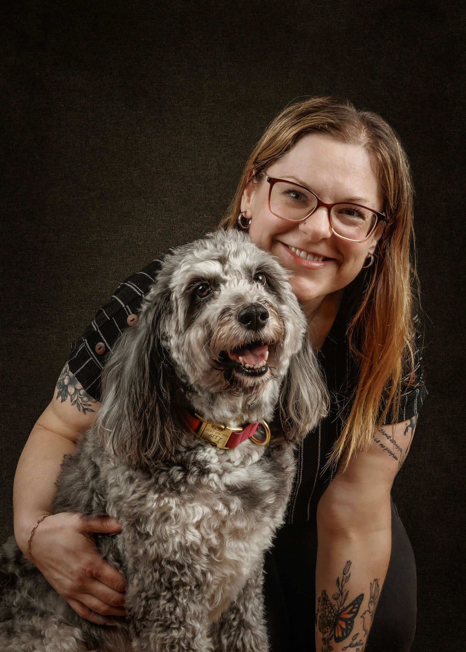 Smiling woman with glasses and tattoos poses with a fluffy gray and white dog wearing a red collar.