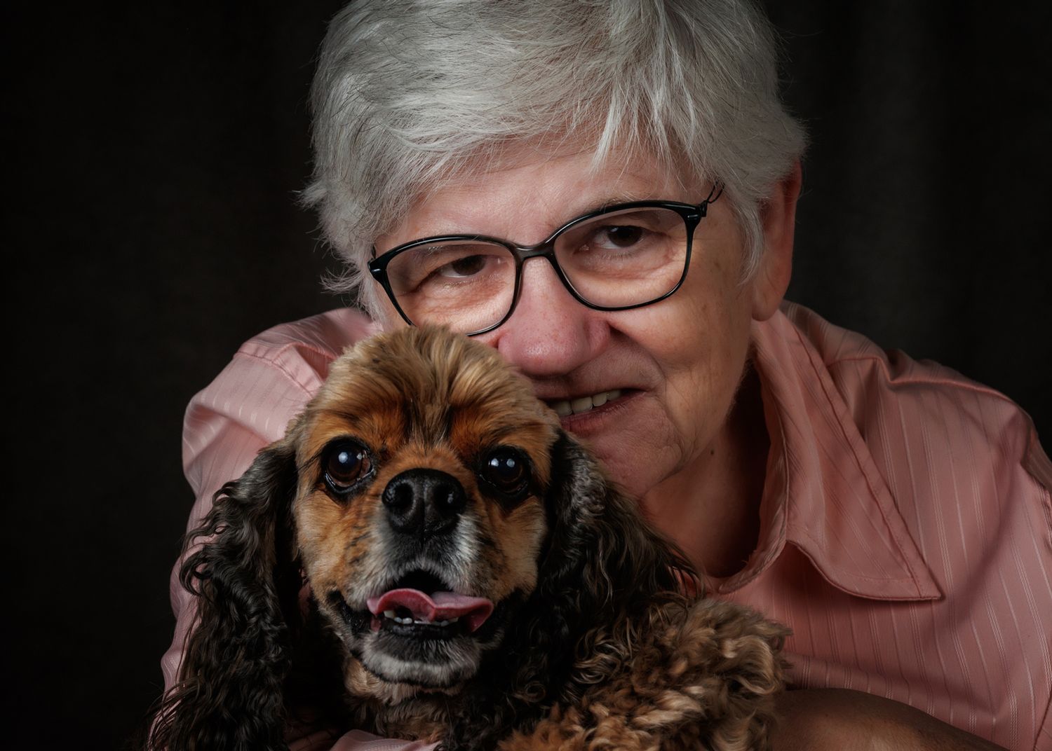 Elderly woman with white hair and glasses smiling while hugging a brown Cocker Spaniel dog against dark background.