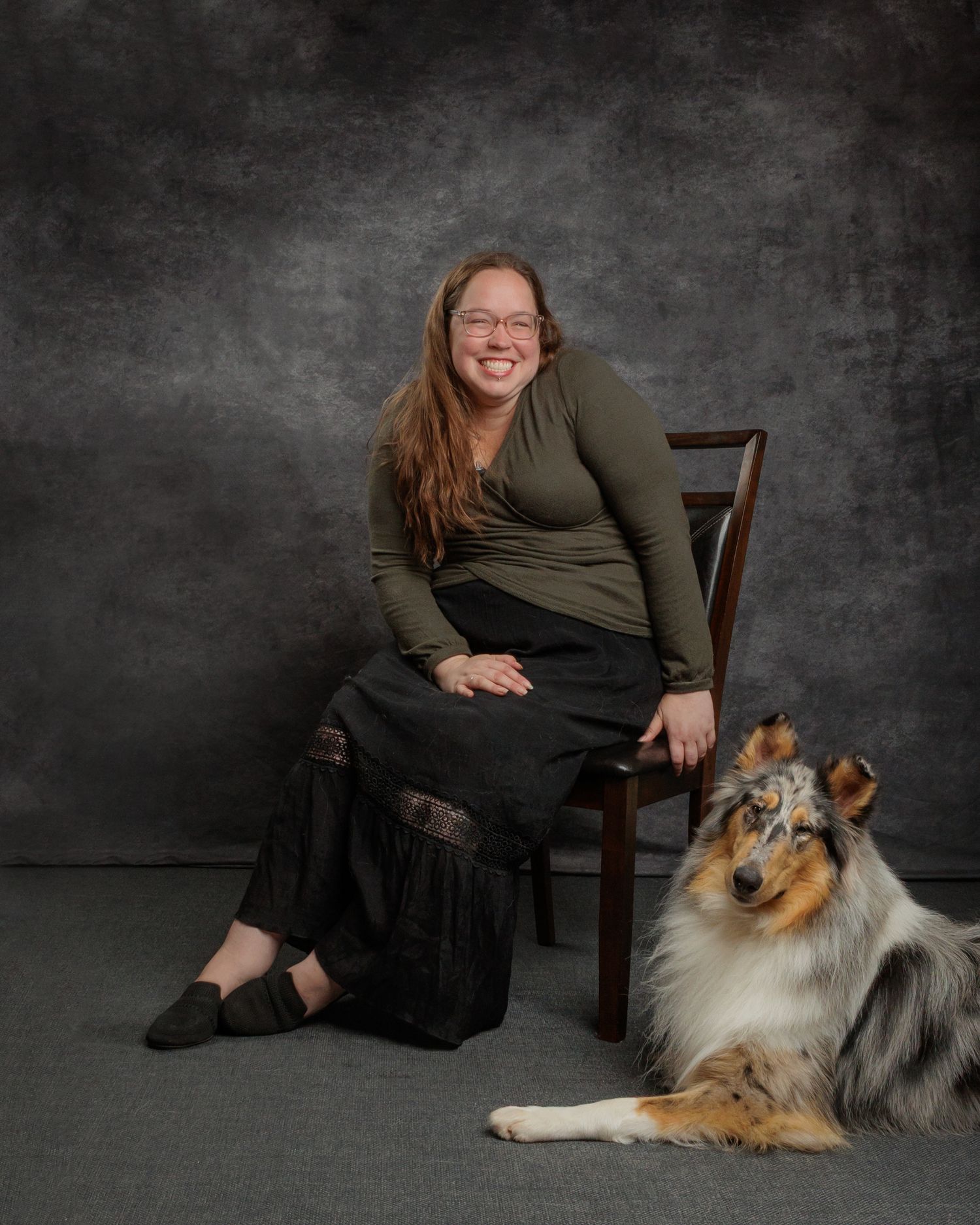 Woman in green top and black skirt sits on chair beside a tri-color Rough Collie dog in a studio portrait.
