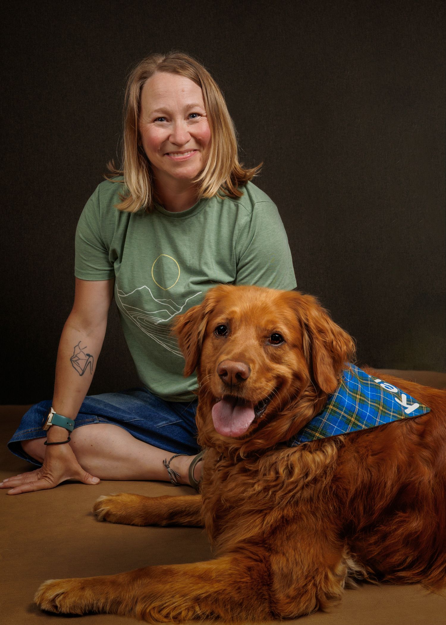 Woman in green t-shirt sitting cross-legged with a golden retriever wearing a blue bandana in a studio portrait.
