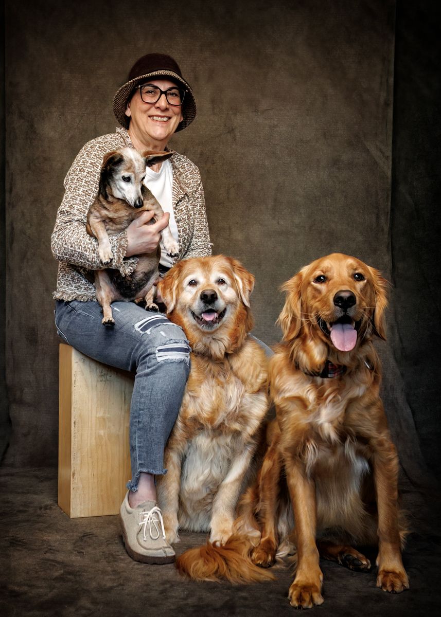 Woman in brown hat holding small dog while seated beside two golden retrievers in a studio portrait.