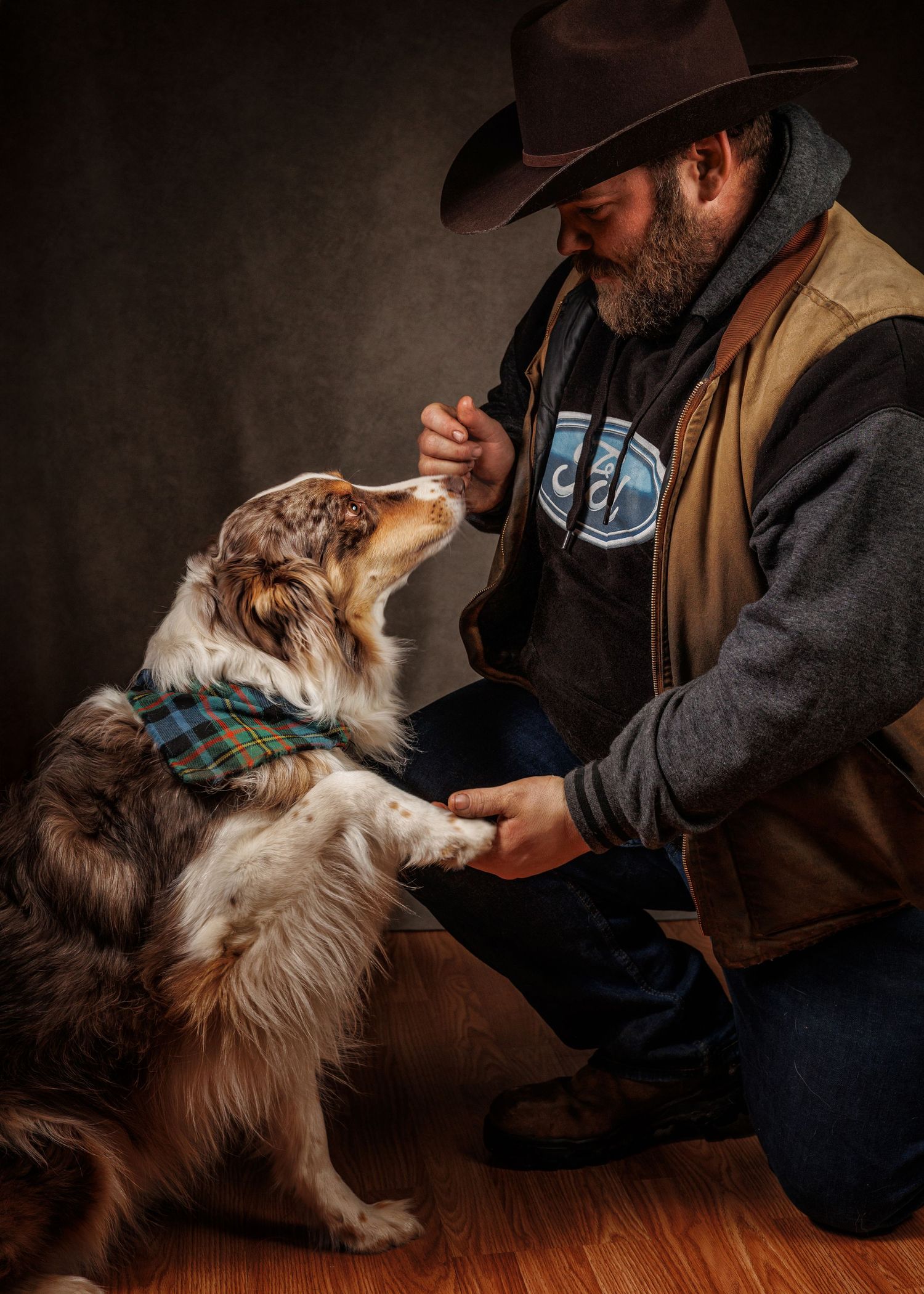 Bearded cowboy in a hat feeds a treat to an Australian Shepherd dog wearing a plaid bandana indoors.