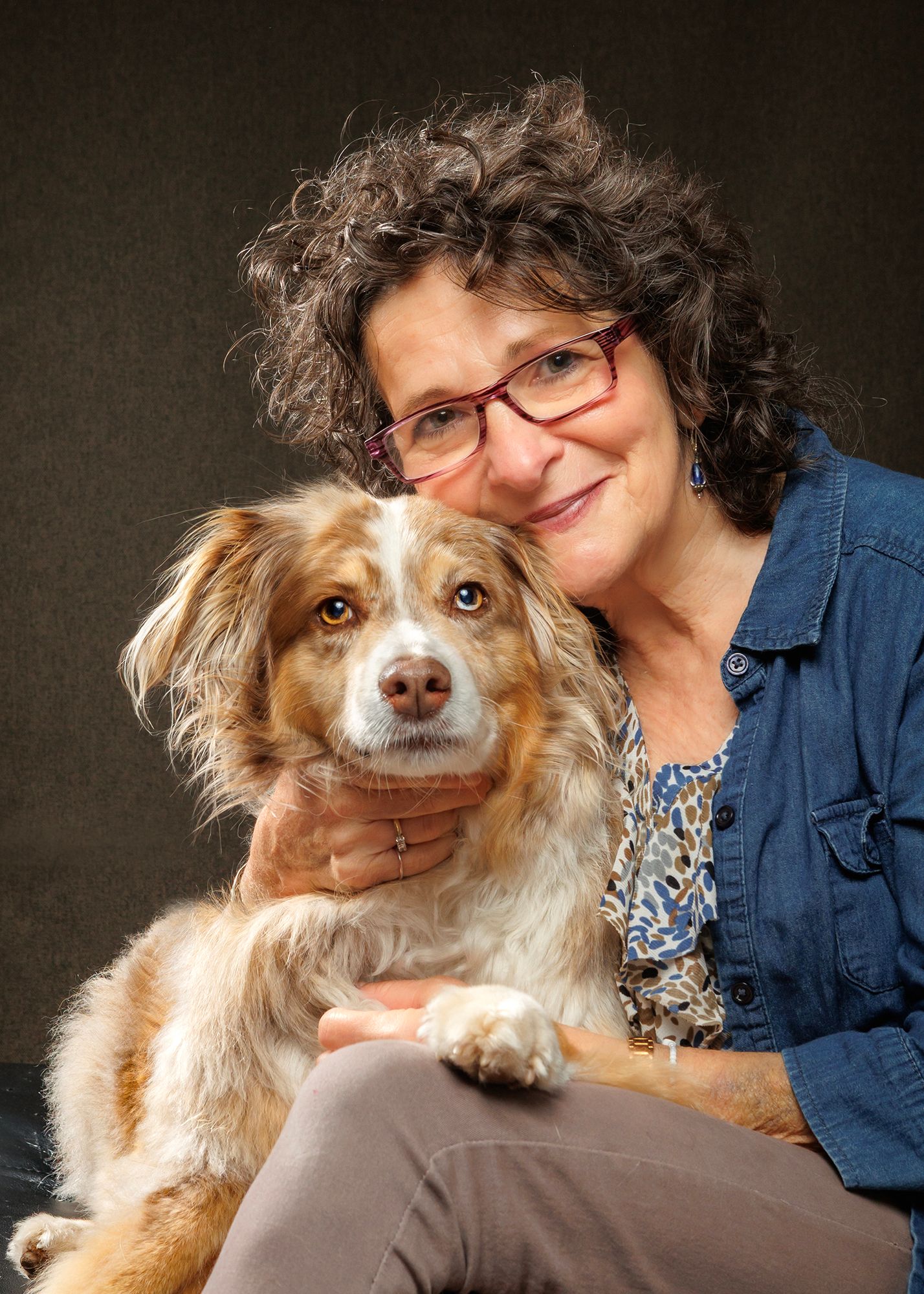 Woman with curly hair and red glasses smiling while hugging a fluffy brown and white Australian Shepherd dog.