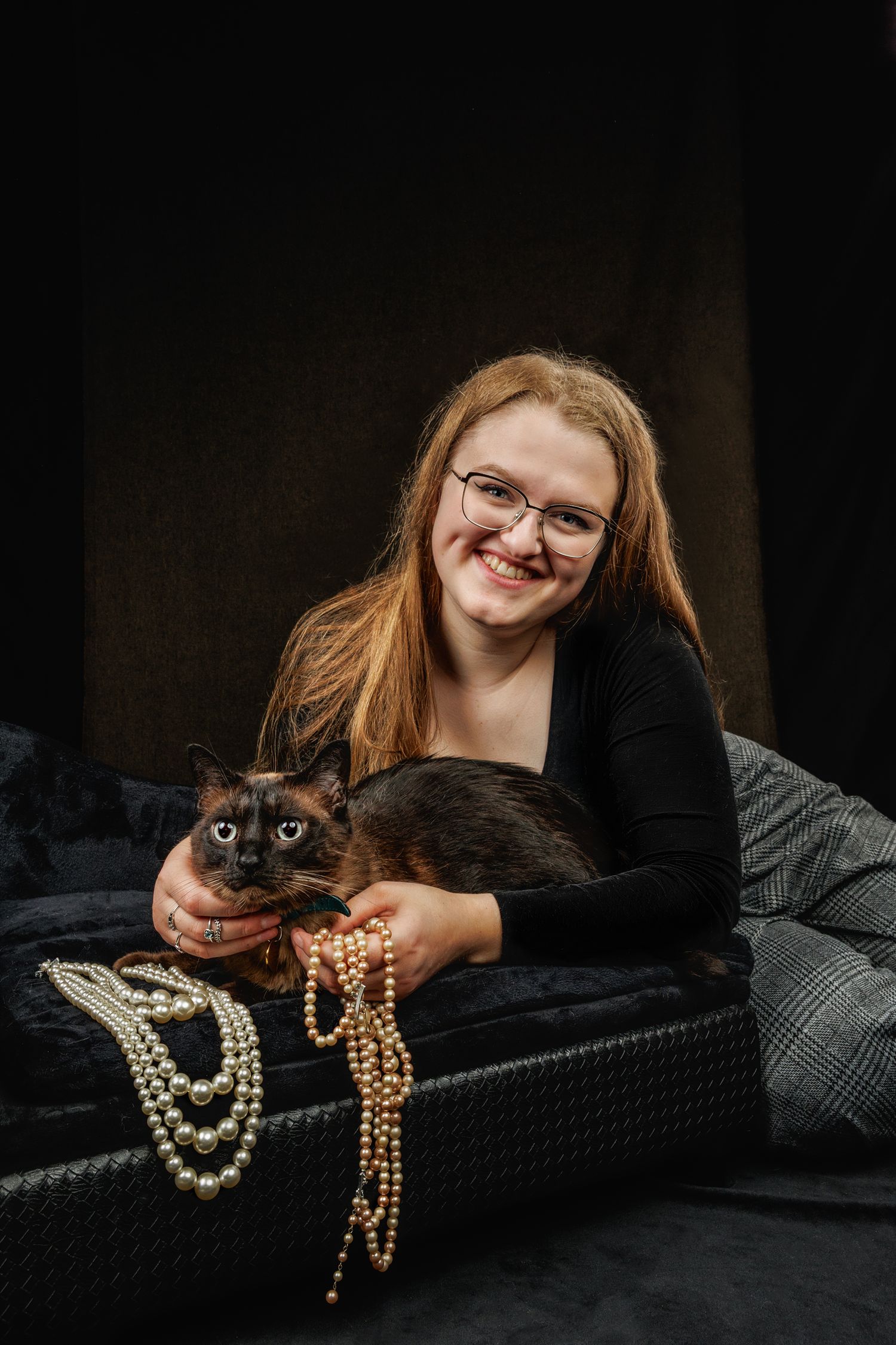 Young woman with red hair and glasses smiles while holding a dark cat, surrounded by pearl necklaces on a black surface.