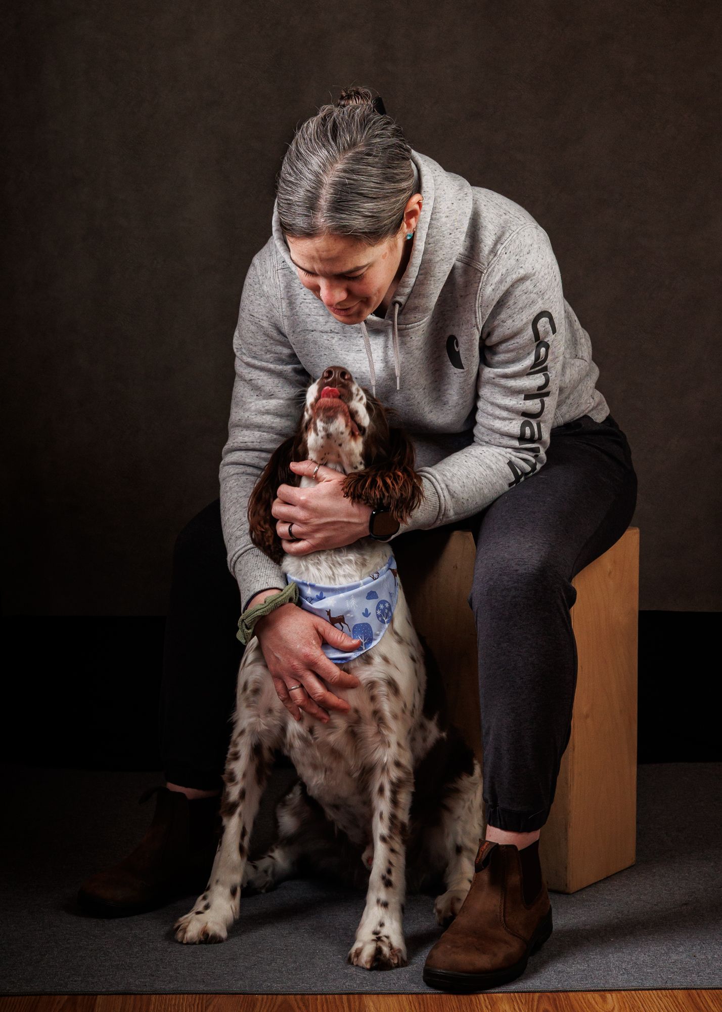 Woman in gray Carhartt sweatshirt sitting with a spotted Springer Spaniel dog in a studio portrait.
