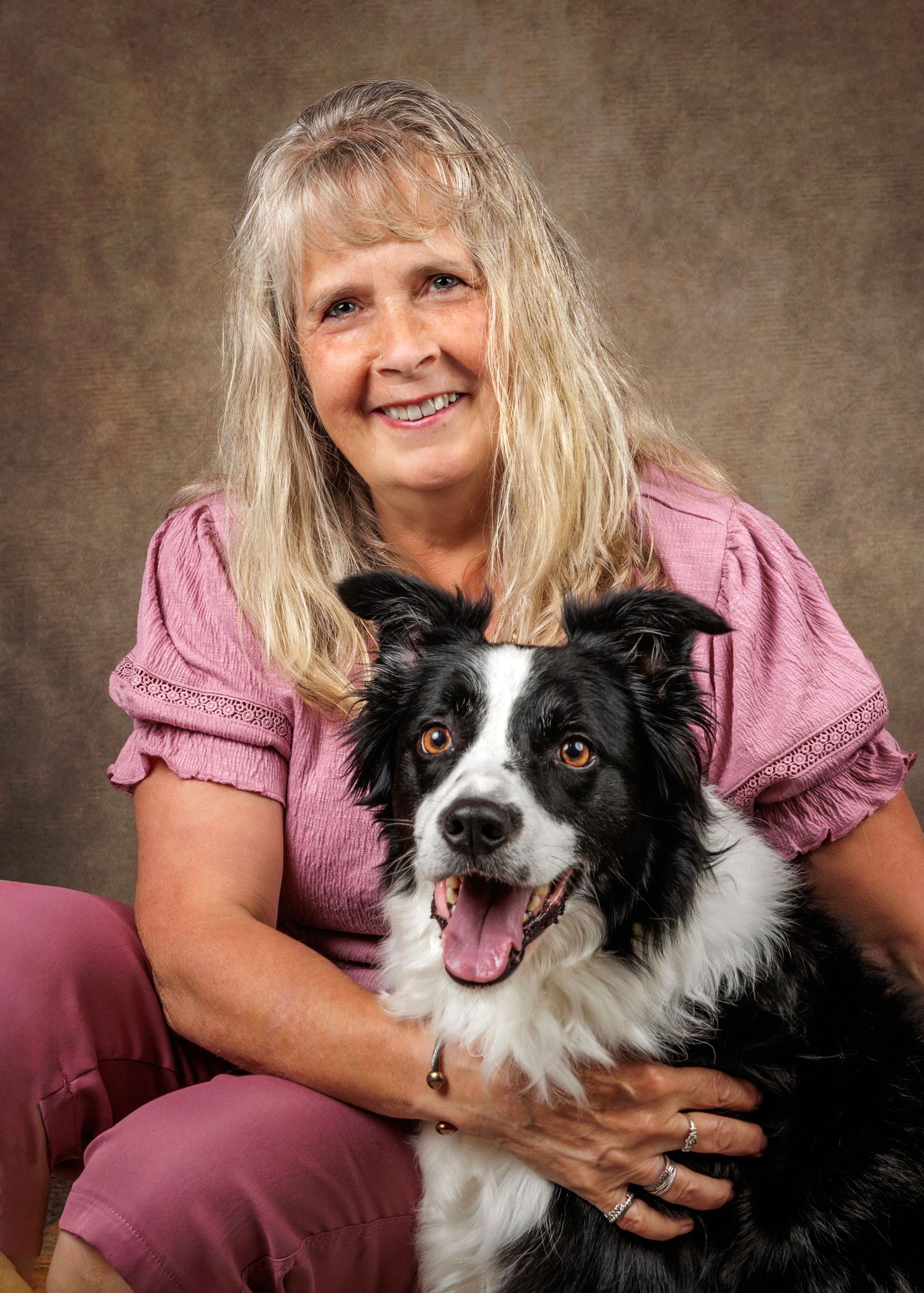 Smiling woman with blonde hair in pink top posing with a black and white Border Collie dog in studio portrait.
