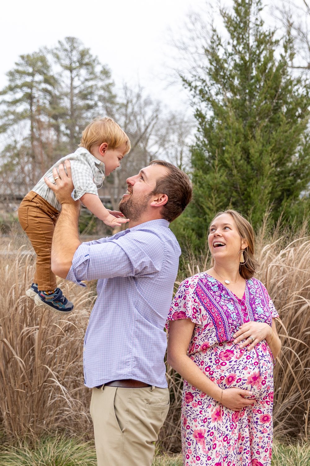 A happy family outdoors, father lifting toddler son while pregnant mother smiles, surrounded by tall grasses and evergreen trees.