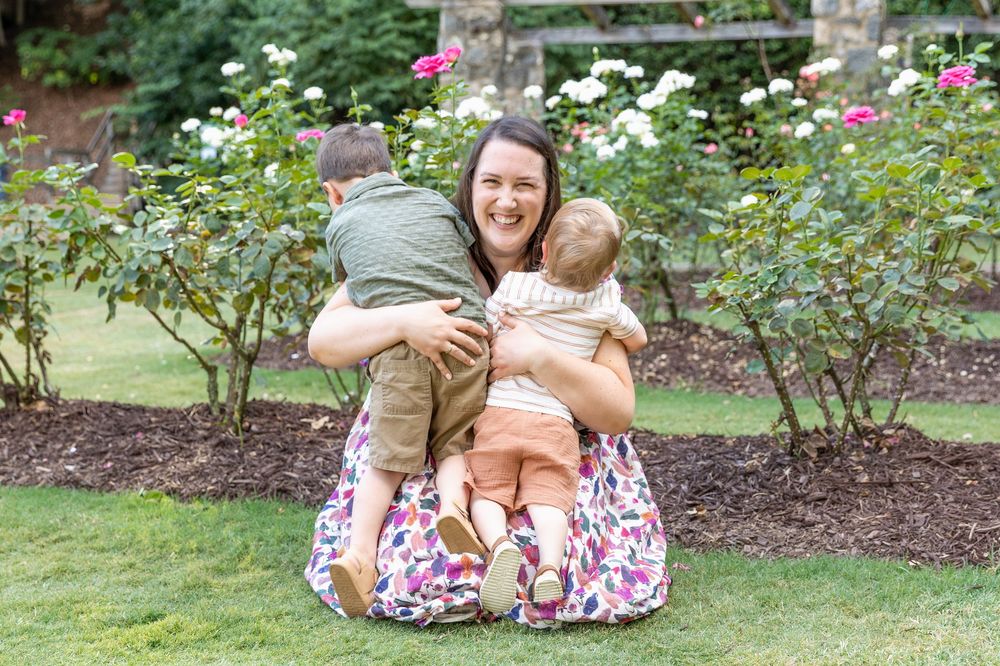 A smiling woman sits on grass hugging two young children in a lush garden with blooming white and pink roses.
