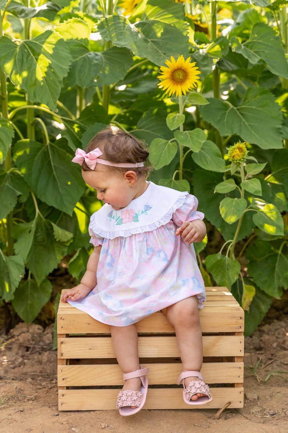 Adorable baby girl in pink floral dress sitting on wooden crate in sunflower garden.