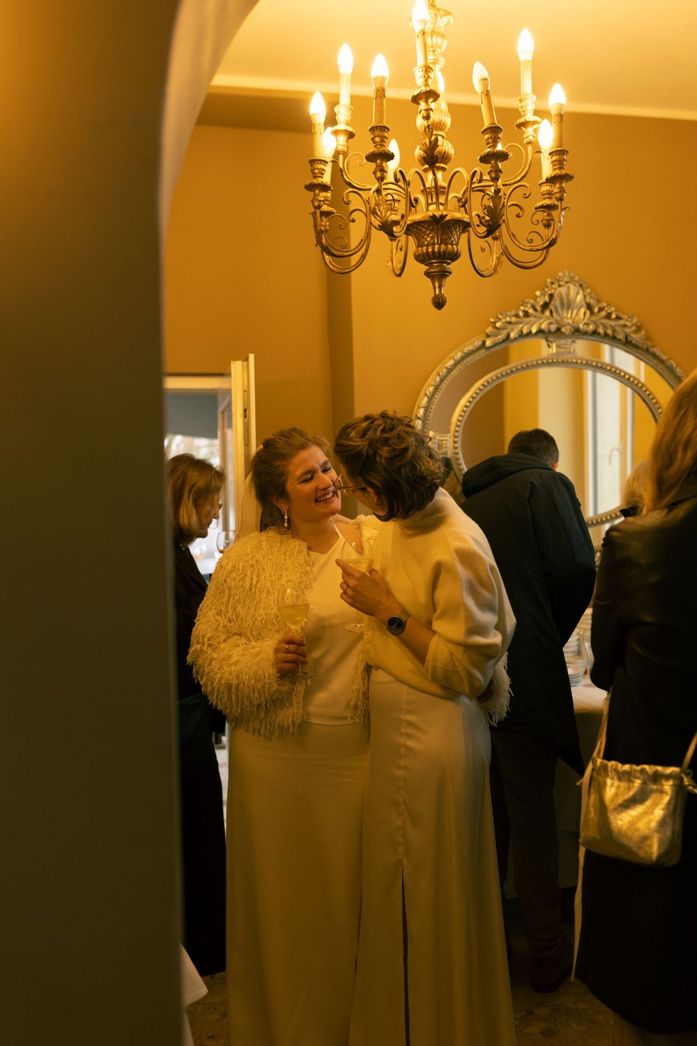 Two women in elegant white dresses embrace and smile beneath a golden chandelier at a formal indoor gathering.