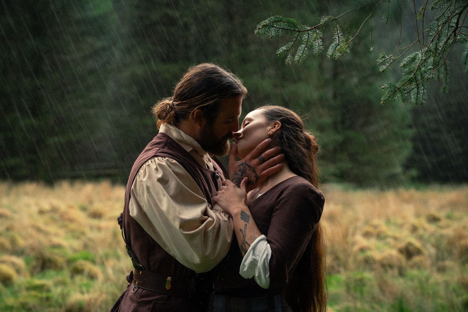 A couple shares a romantic kiss in the rain surrounded by lush green forest in a period drama scene.