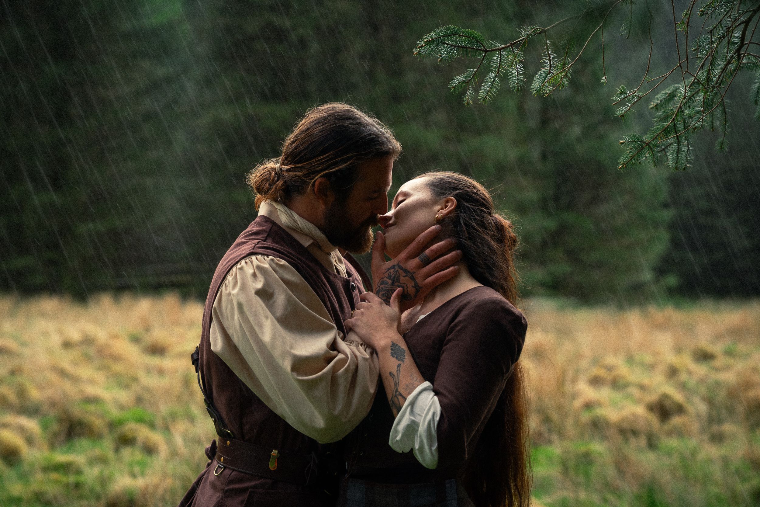 A couple shares a romantic kiss in the rain surrounded by lush green forest in a period drama scene.