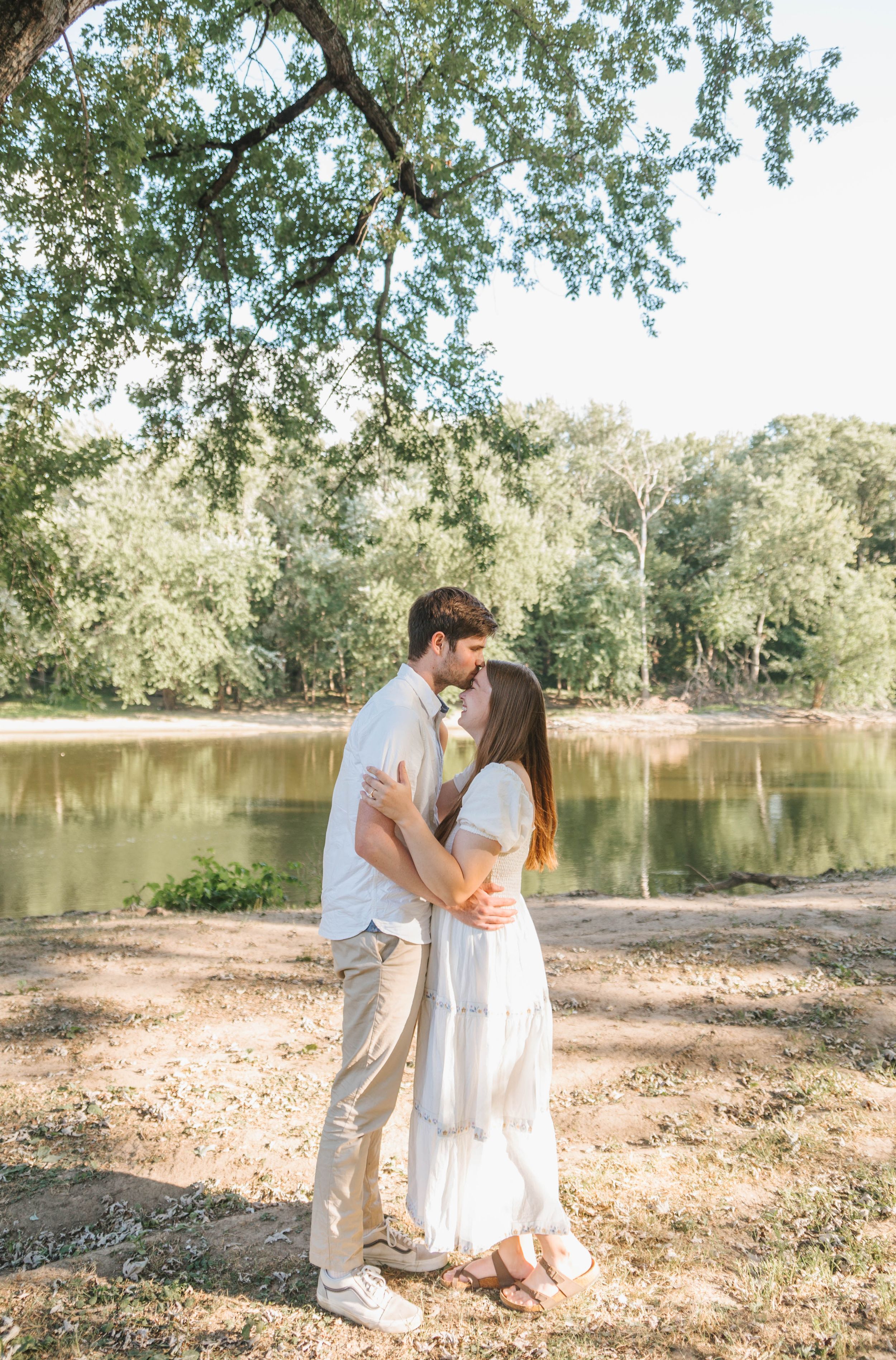 Couple kissing near a serene lake under a large tree, wearing white outfits in a lush green outdoor setting.