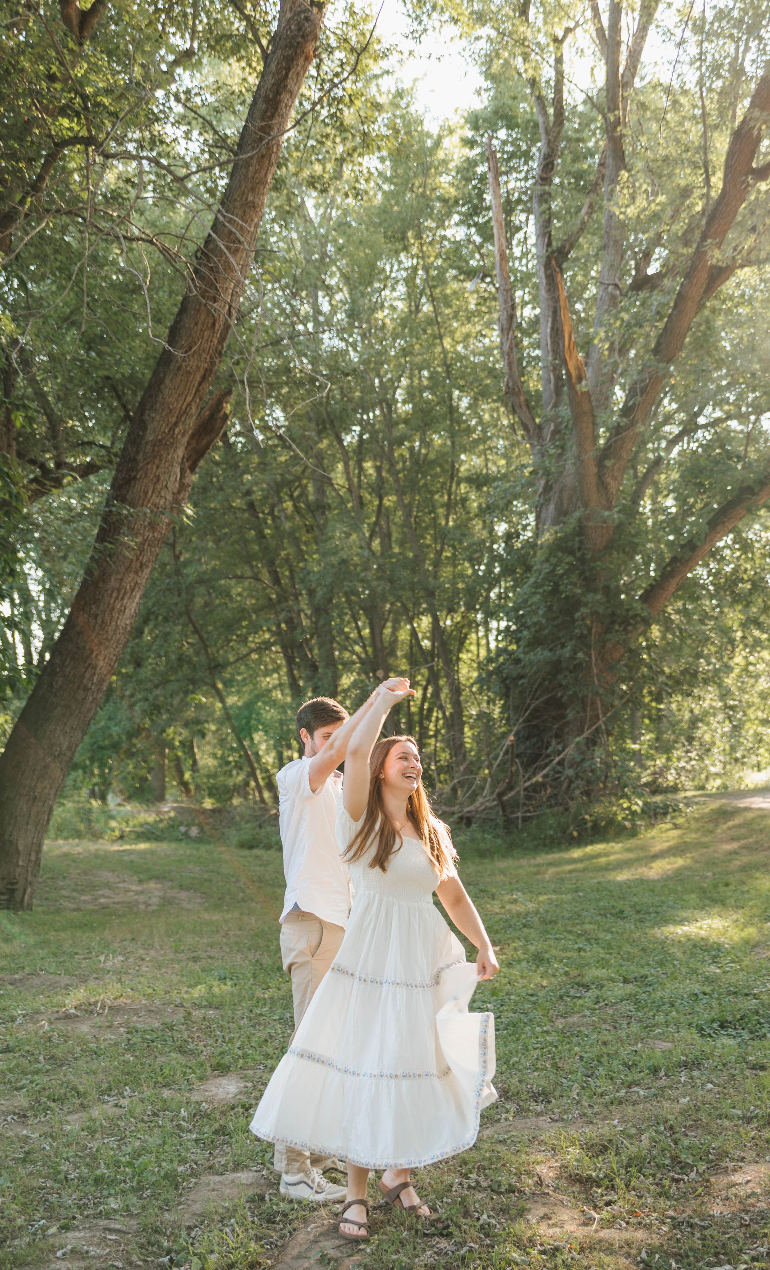 A couple dances outdoors in a sunlit forest, woman in white tiered dress being twirled by her partner.