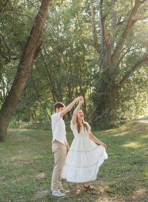 A couple dances joyfully in a sunlit forest, the woman twirling in a white dress while her partner spins her around.