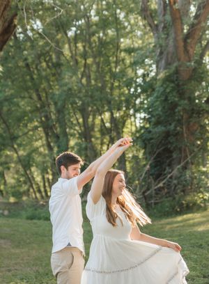 A couple dances joyfully outdoors in a lush green park, the woman in a flowing white dress being twirled by her partner.