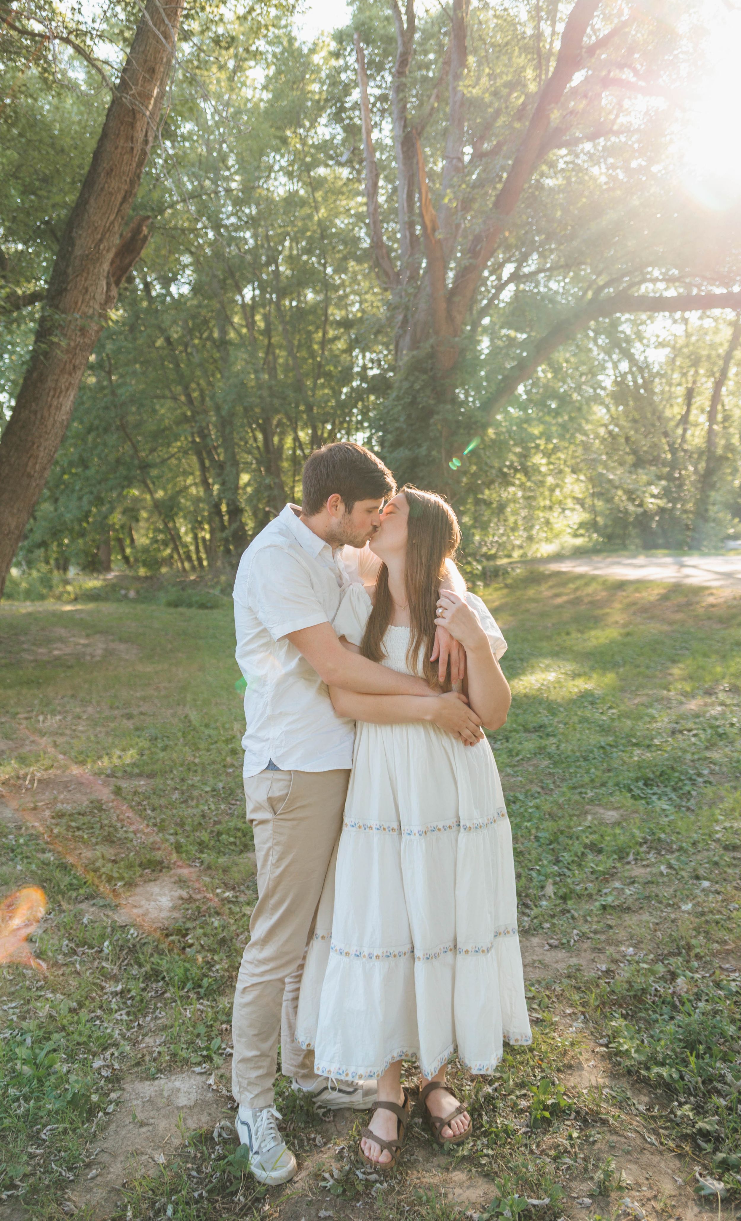 A couple shares a romantic kiss outdoors in a sunlit forest, the woman wearing a flowing white dress.