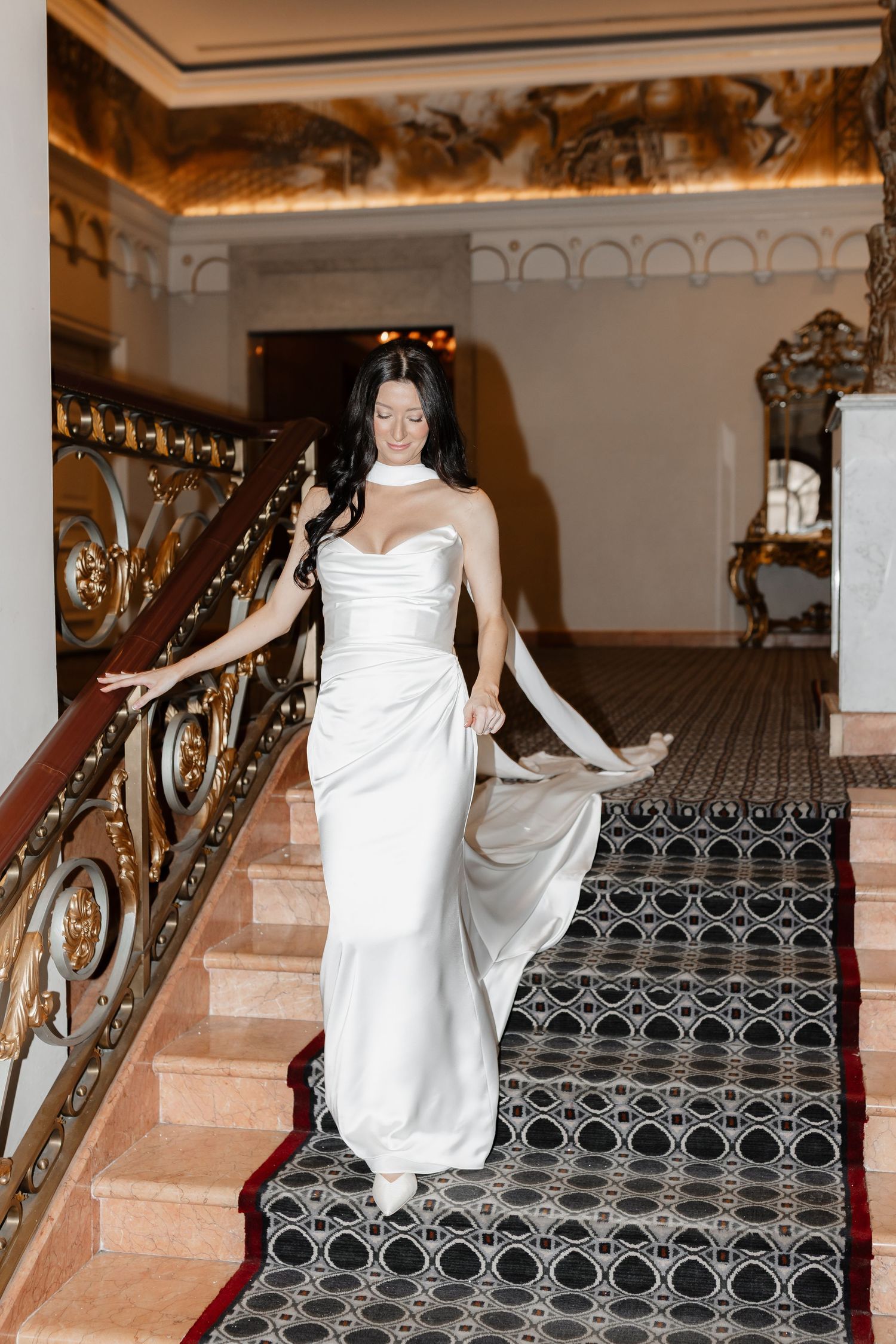 Elegant bride in white satin gown descending ornate staircase with gold railings in a luxurious venue.