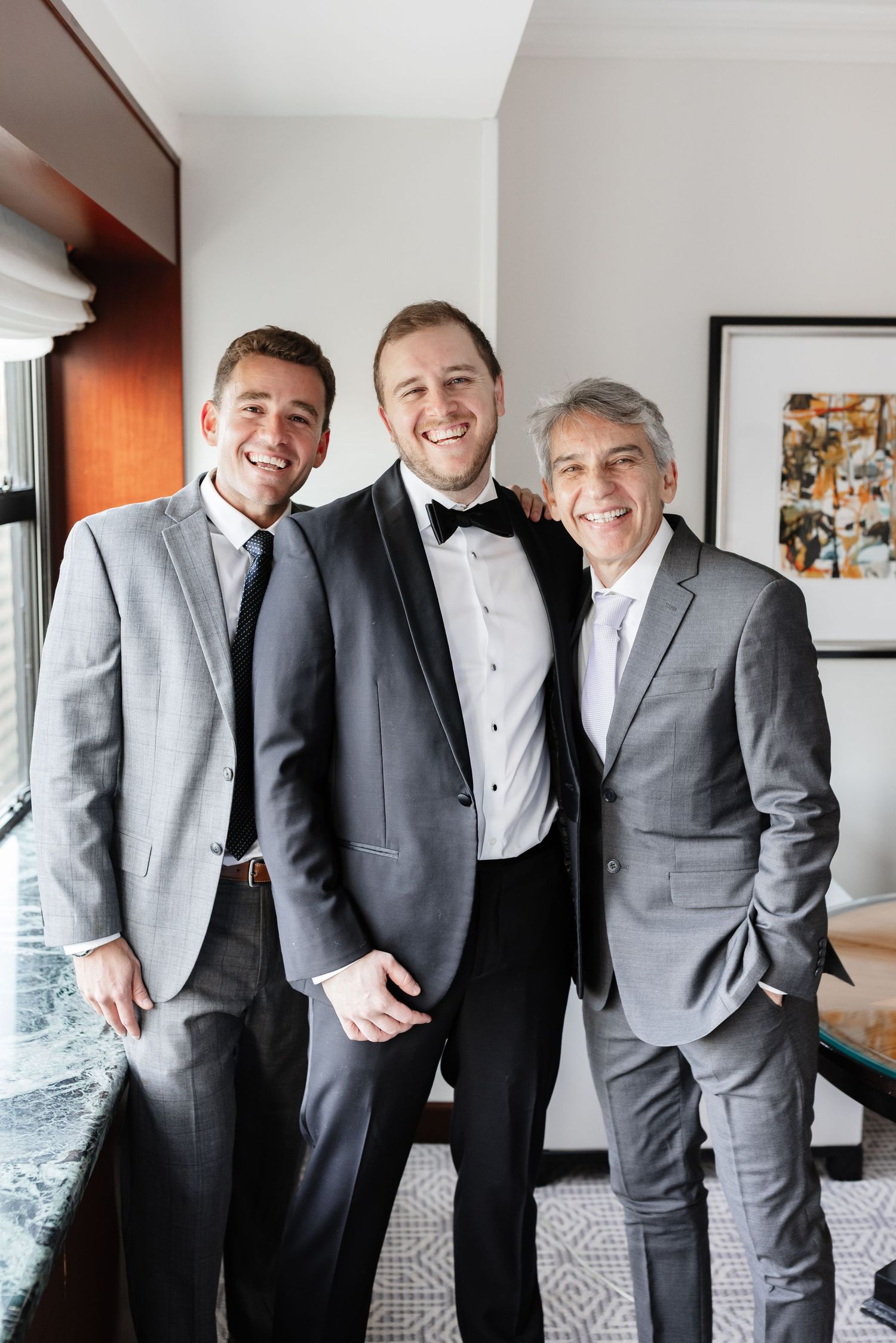 Three men in formal suits smile together indoors, with the center man wearing a tuxedo, likely at a wedding celebration.