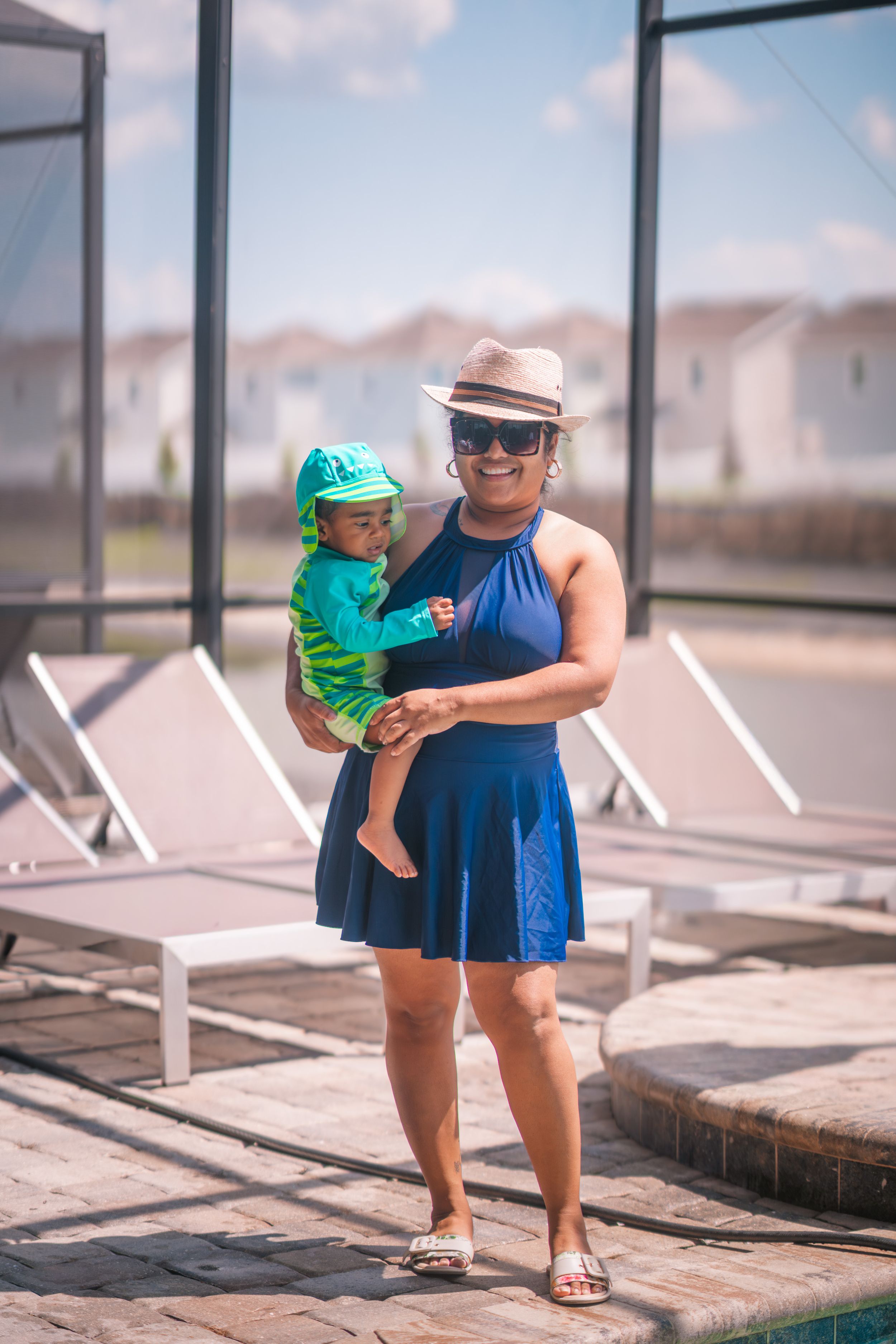 Woman in blue dress and hat holds baby in green outfit poolside on sunny day.