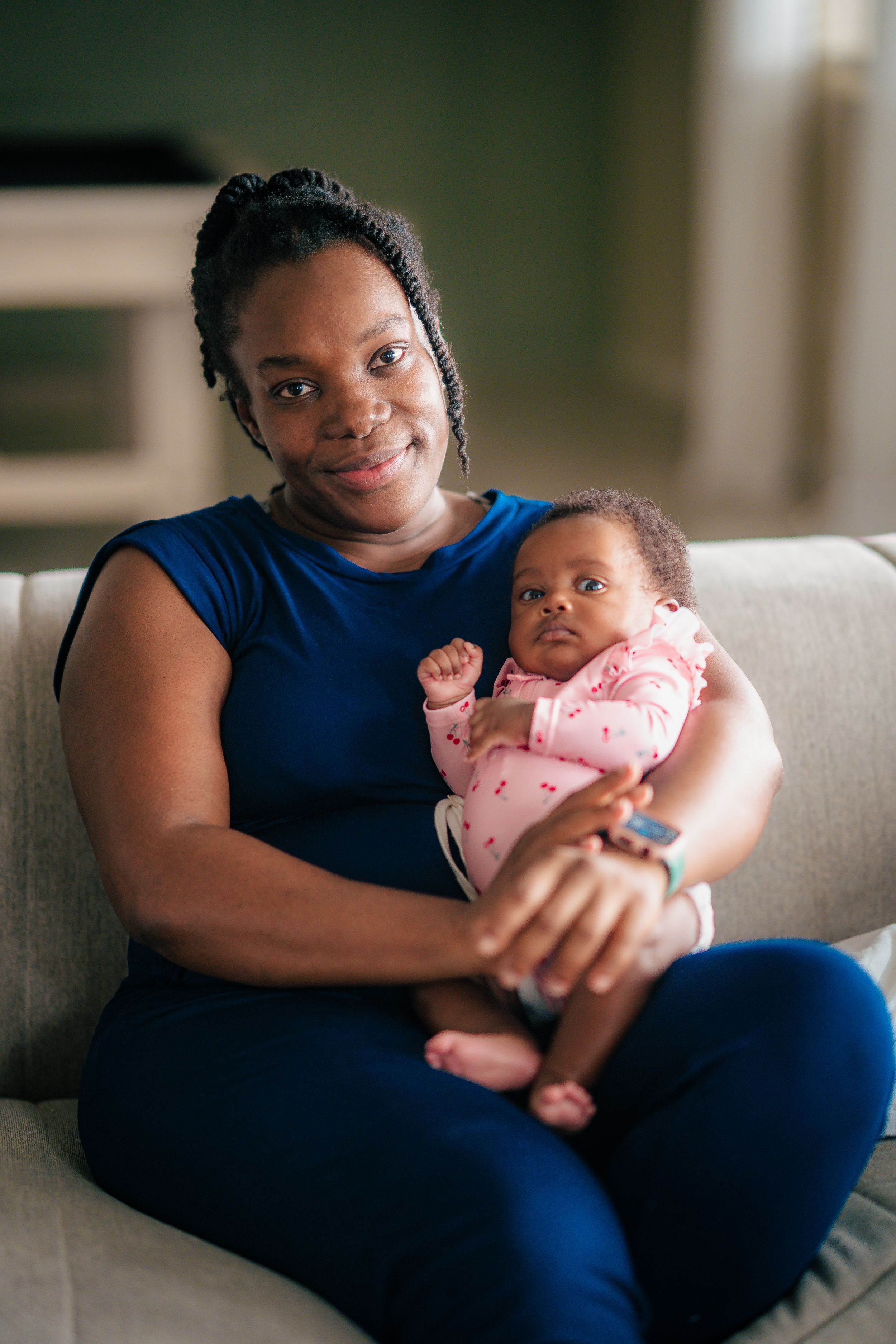 A smiling woman in blue holds her newborn baby in pink while sitting on a couch at home.