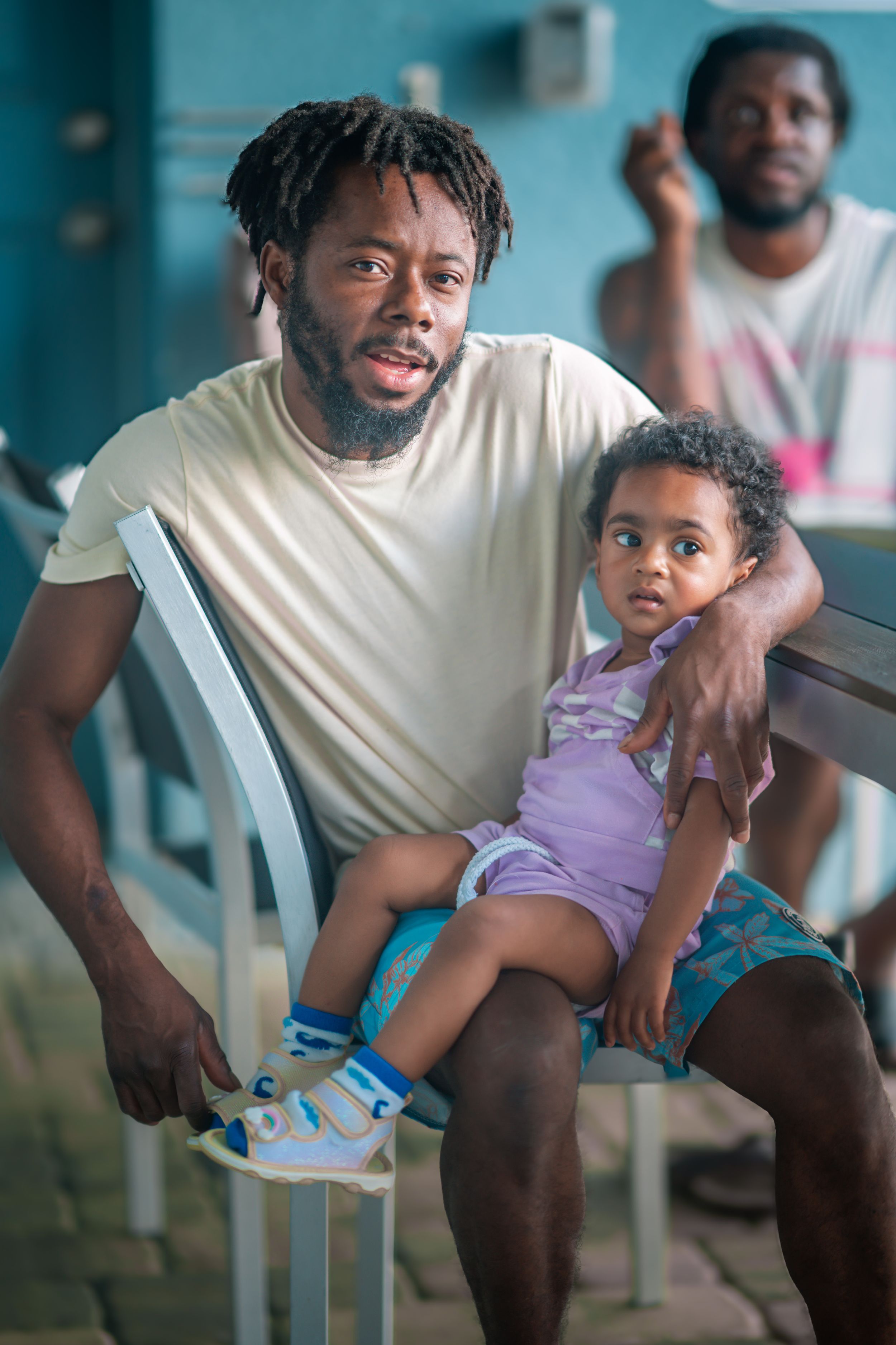 A man with dreadlocks sits on a chair holding a young child on his lap, with another man visible in the background.
