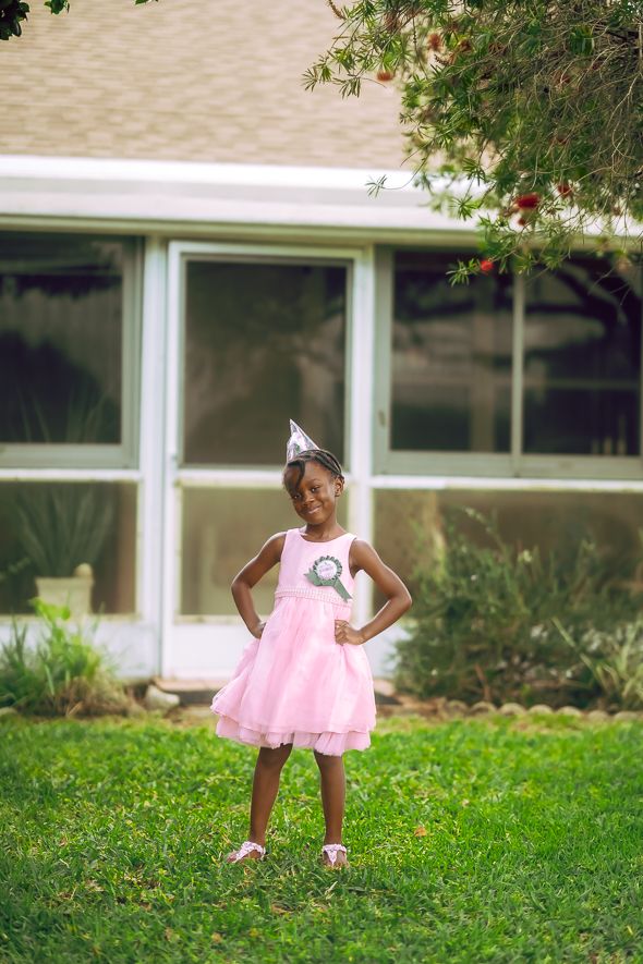 A young Black girl in a pink dress and party hat stands confidently on green grass in front of a house.