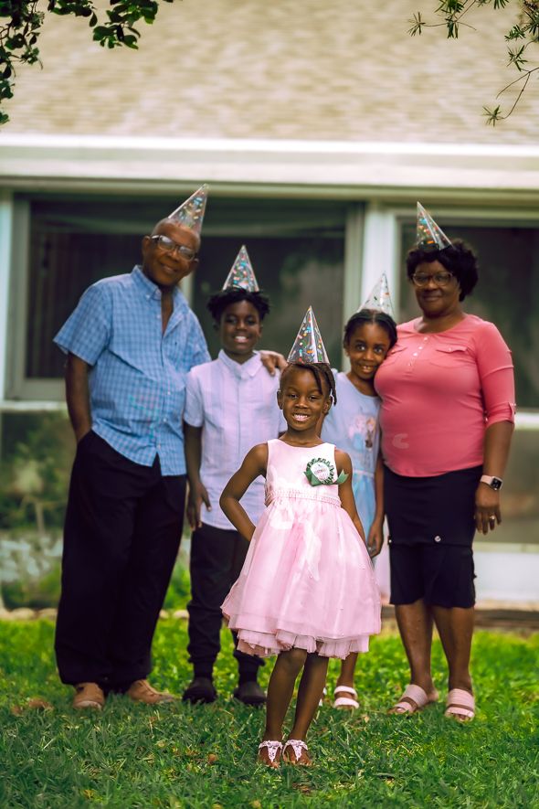 Family of five wearing party hats celebrates a birthday outdoors, with young girl in pink dress as the guest of honor.