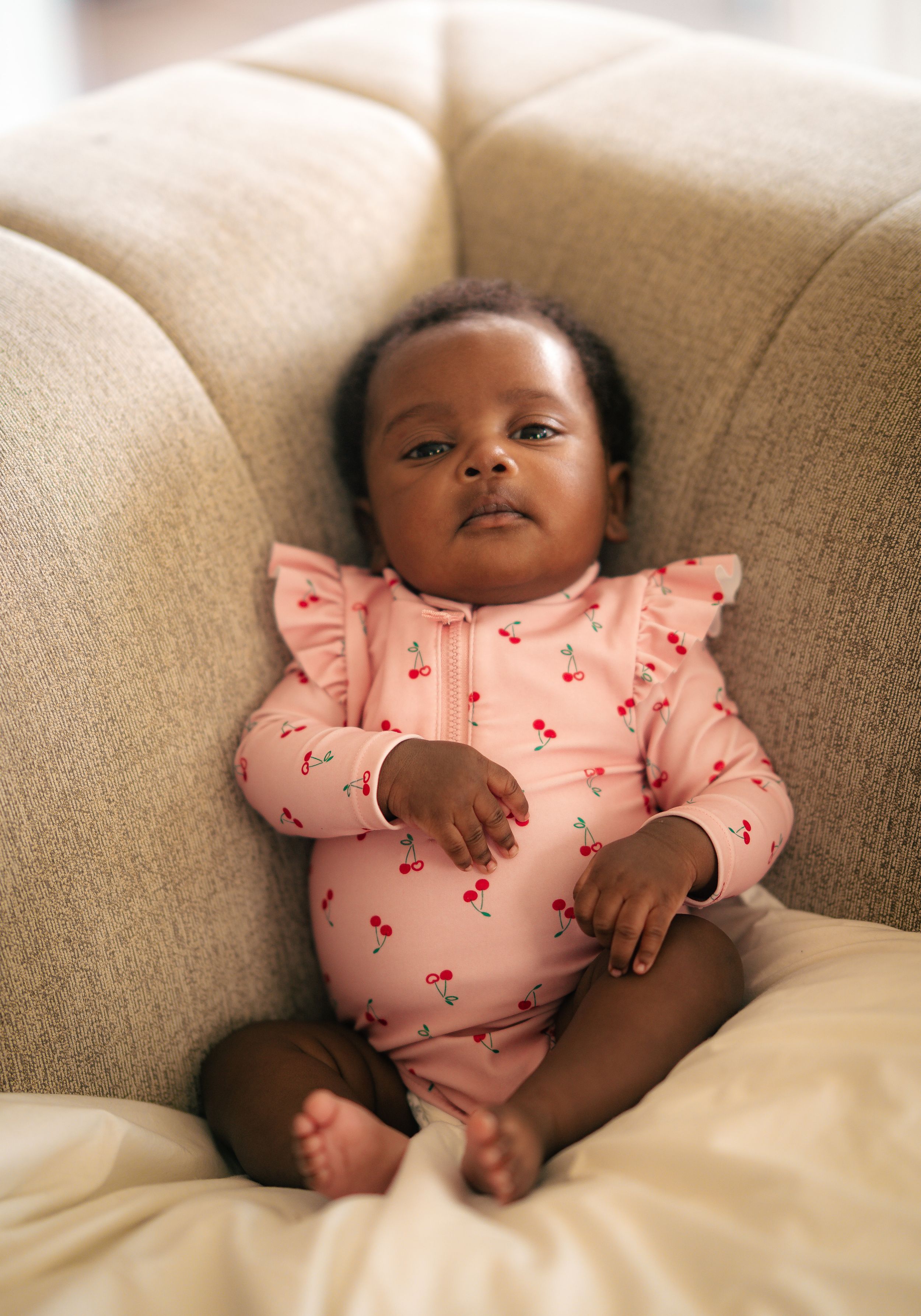 Adorable baby girl in pink cherry-print ruffle romper sitting on a beige sofa, looking at the camera.