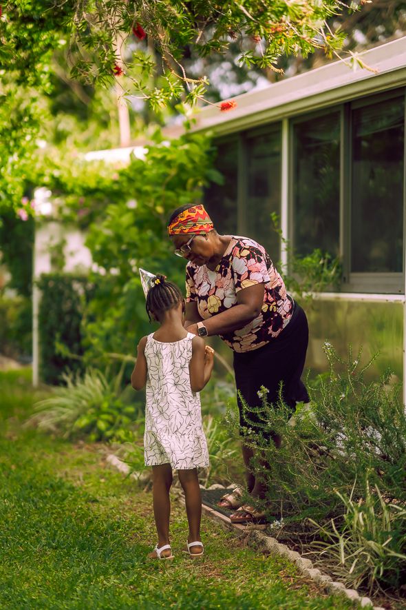 A grandmother in a floral top and headwrap bends to interact with a young girl in a white dress in a lush green garden.