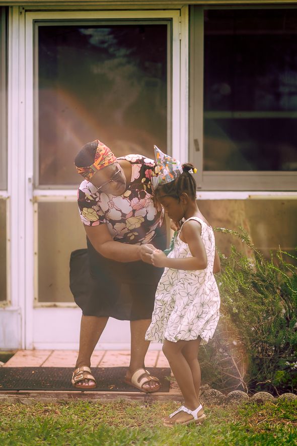 Adult and young girl in floral outfits interact outside a house on a sunny day.