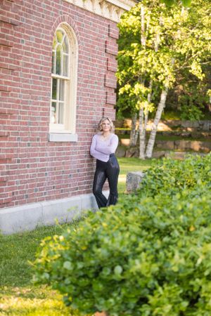 A blonde woman in a lavender top leaning against a red brick building surrounded by green foliage.