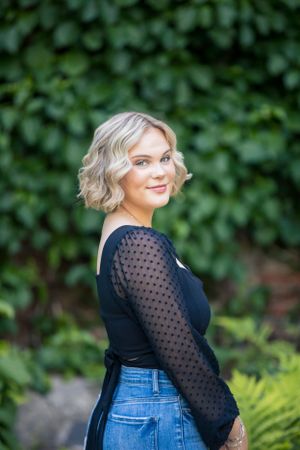 Blonde woman in black polka dot sheer top smiles over shoulder against lush green ivy wall.