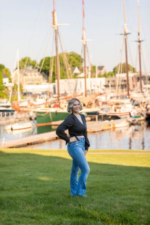 Woman in black top and jeans posing confidently by a marina with sailboats at golden hour.